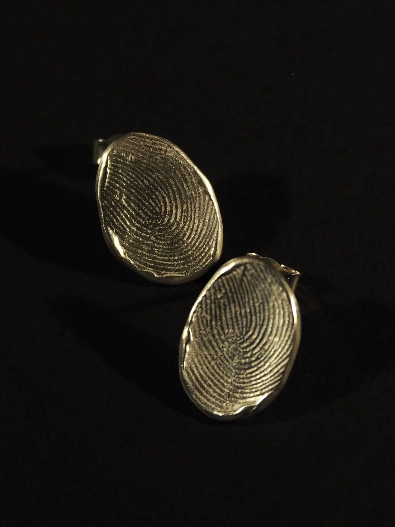 Deux boucles d’oreilles ovales en argent sur fond noir dévoilent un motif d’empreinte digitale. Ces bijoux empreinte symbolisent le lien unique d’une création sur mesure, élégante et moderne, conçue par une artisane lyonnaise.