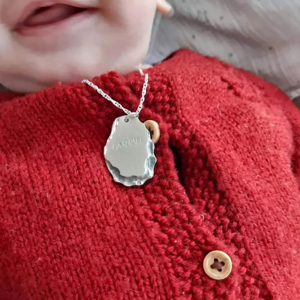 Bébé Gabriel porte son collier de naissance en argent massif gravé de son prénom. Le pendentif aux motifs doux repose sur sa combinaison, création artisanale lyonnaise pour nouveau-né.