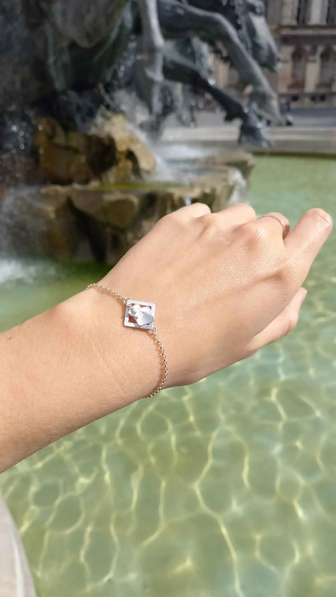 Bracelet pépite fin en argent recyclé au poignet d’une jeune femme devant la Fontaine des Terreaux à Lyon. Reflets aquatiques subliment cette pièce artisanale locale au style minimaliste et contemporain.