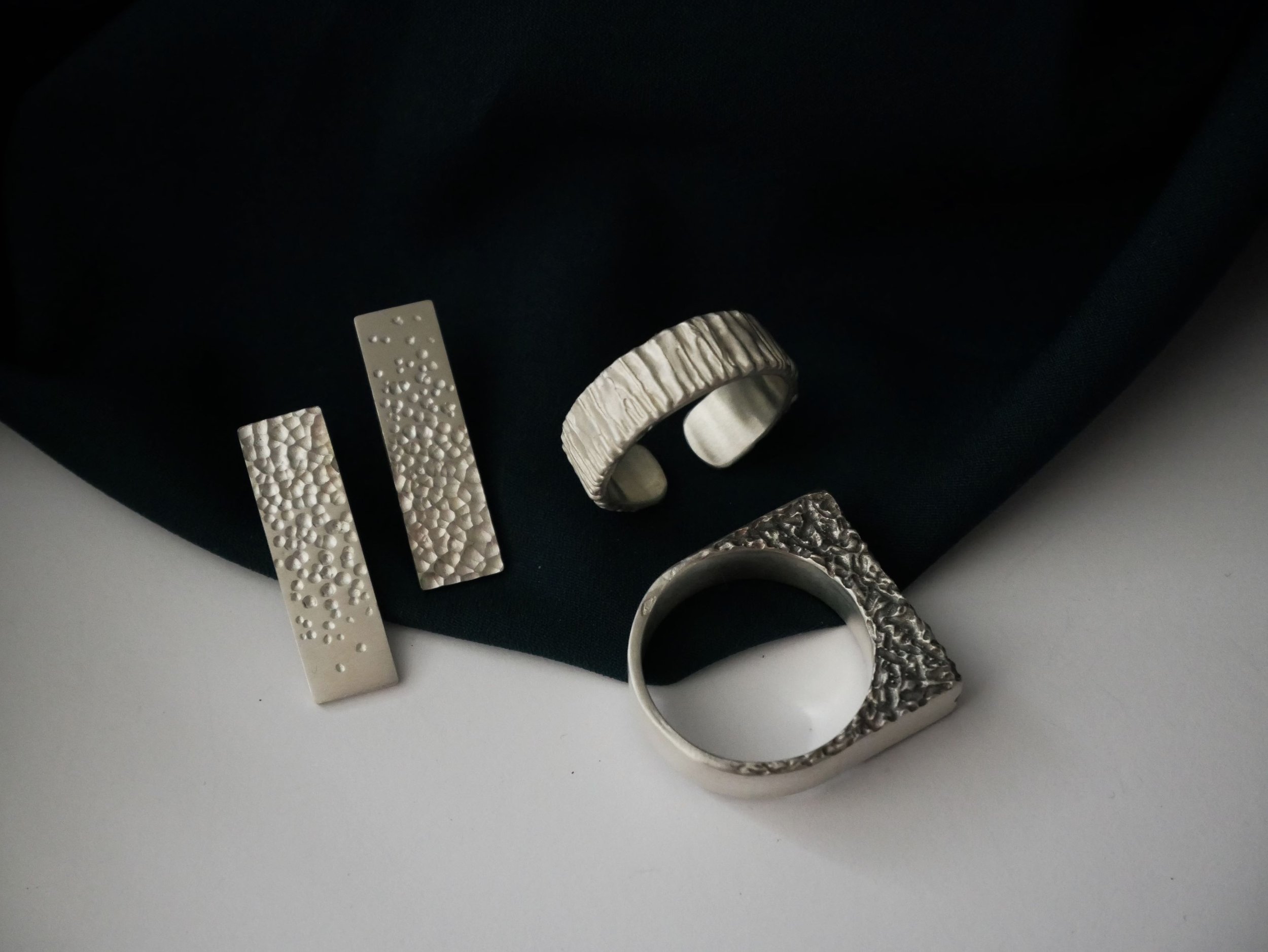 Plusieurs bijoux en argent : boucles d’oreilles rectangulaires texturées, bague ouverte et bague carrée texturée, sur tissu noir fond blanc. Collection harmonieuse de bagues en argent.