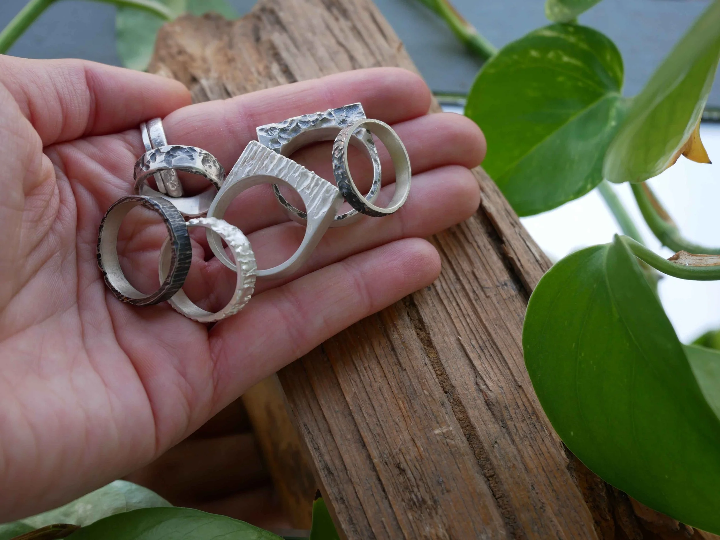 Bague en argent faite main tenue par la bijoutière, avec plante en arrière-plan : anneaux et carrées aux jeux de textures. Pièces artisanales uniques en argent 925 jouant sur reliefs pour un style personnel.