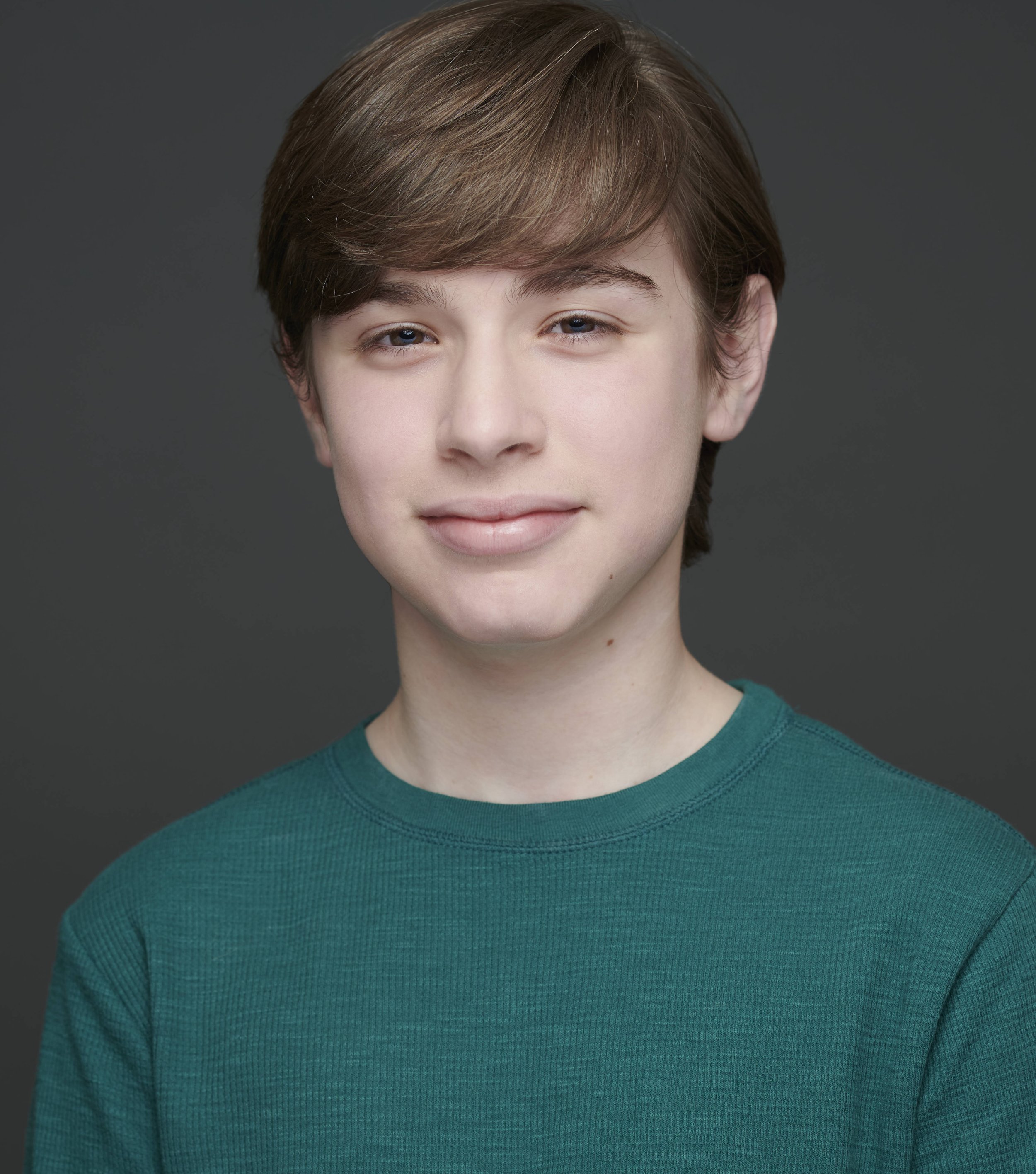 A young person with short, light brown hair wearing a green shirt, smiling softly in front of a gray background.