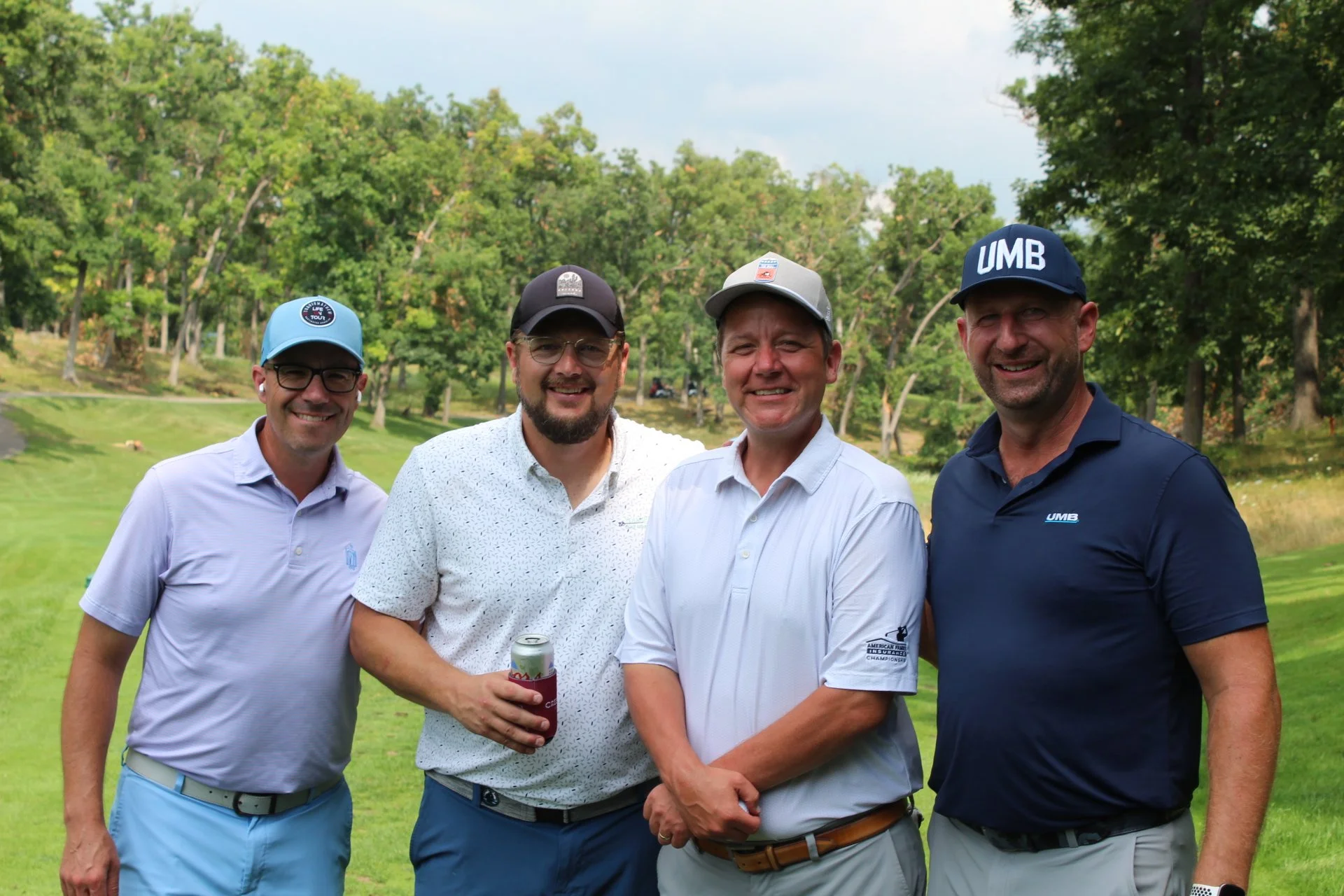 Four men smiling outdoors on a golf course, wearing golf hats and casual polo shirts, with trees in the background.