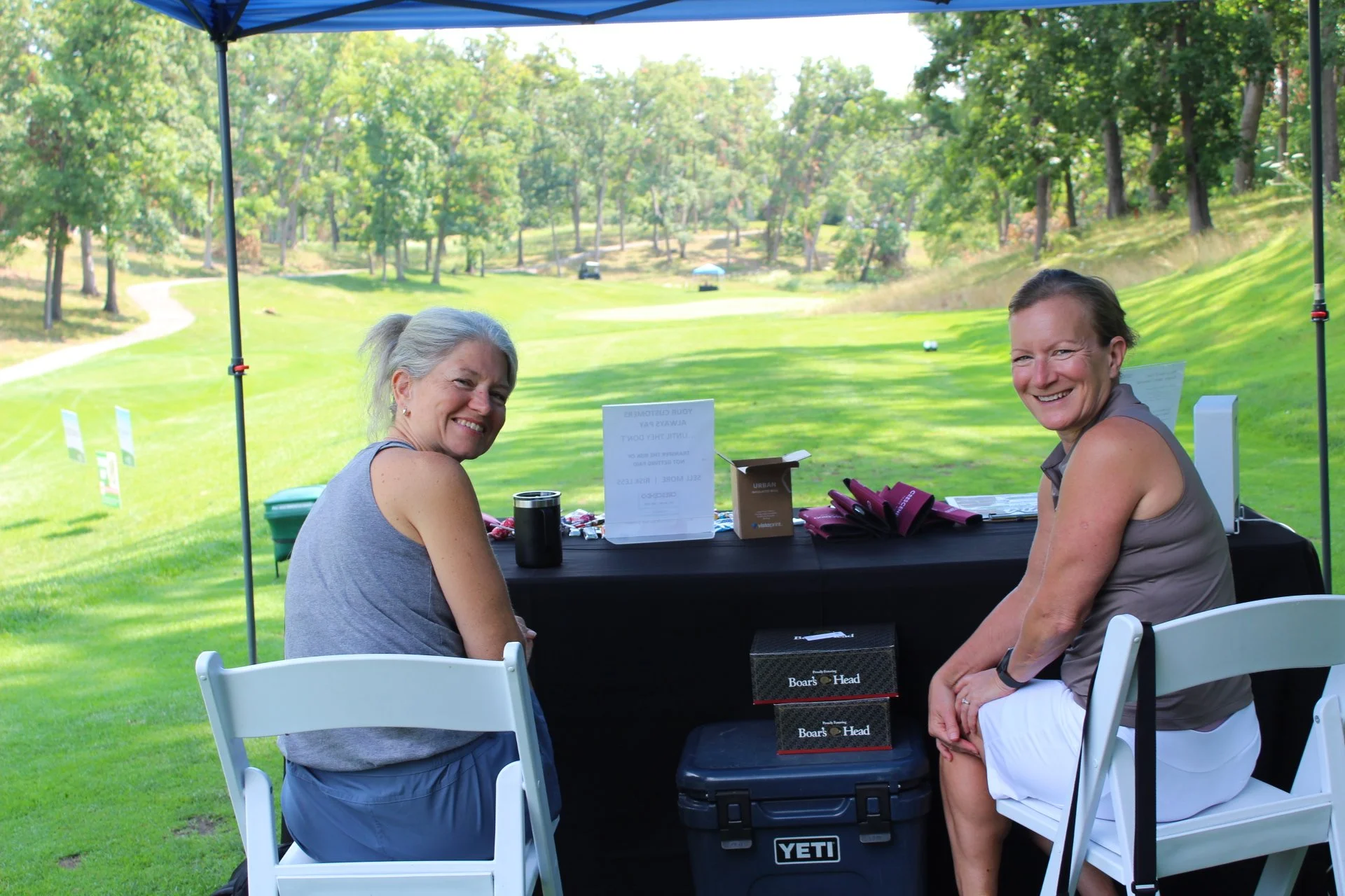 Two women sitting at a table under a blue canopy on a sunny day. They are smiling and looking at the camera. The background features a grassy golf course with trees.