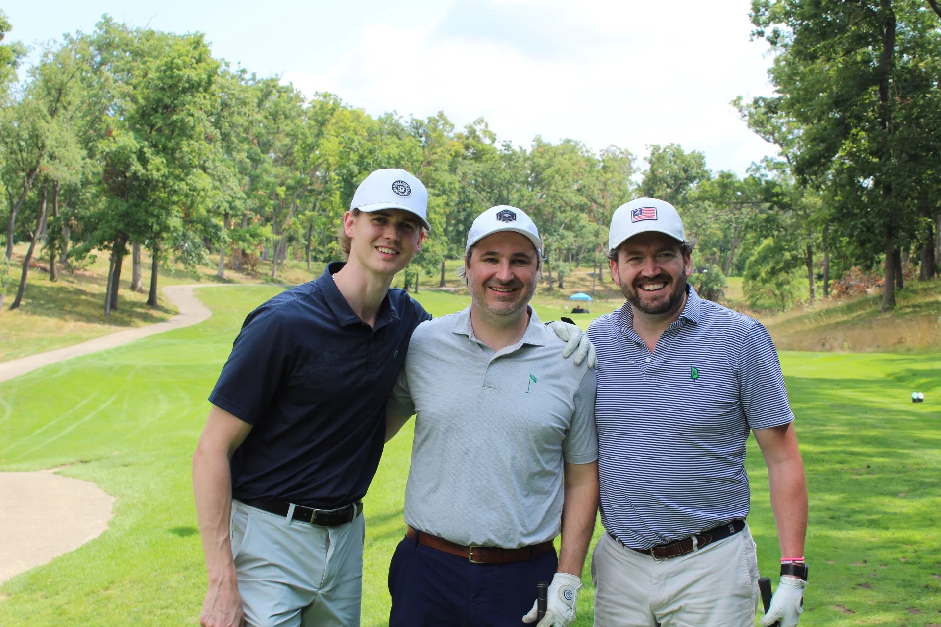 Three men standing on a golf course, smiling and posing for a photo. They are wearing golf caps and casual golf attire, with golf gloves in hand. The background features lush green trees and a slightly overcast sky.