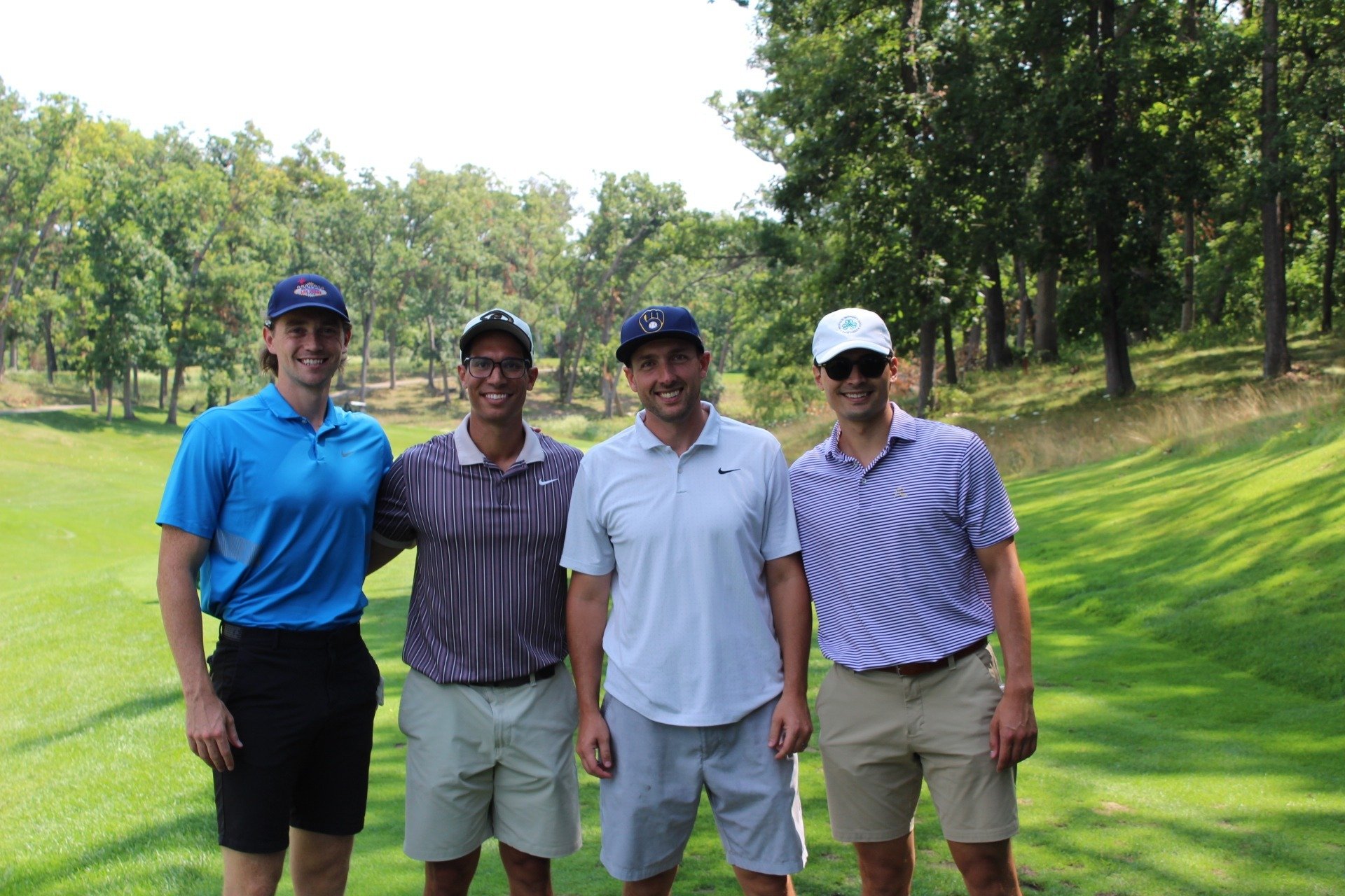 Four men standing outdoors on a golf course, smiling, with trees and green grass in the background.