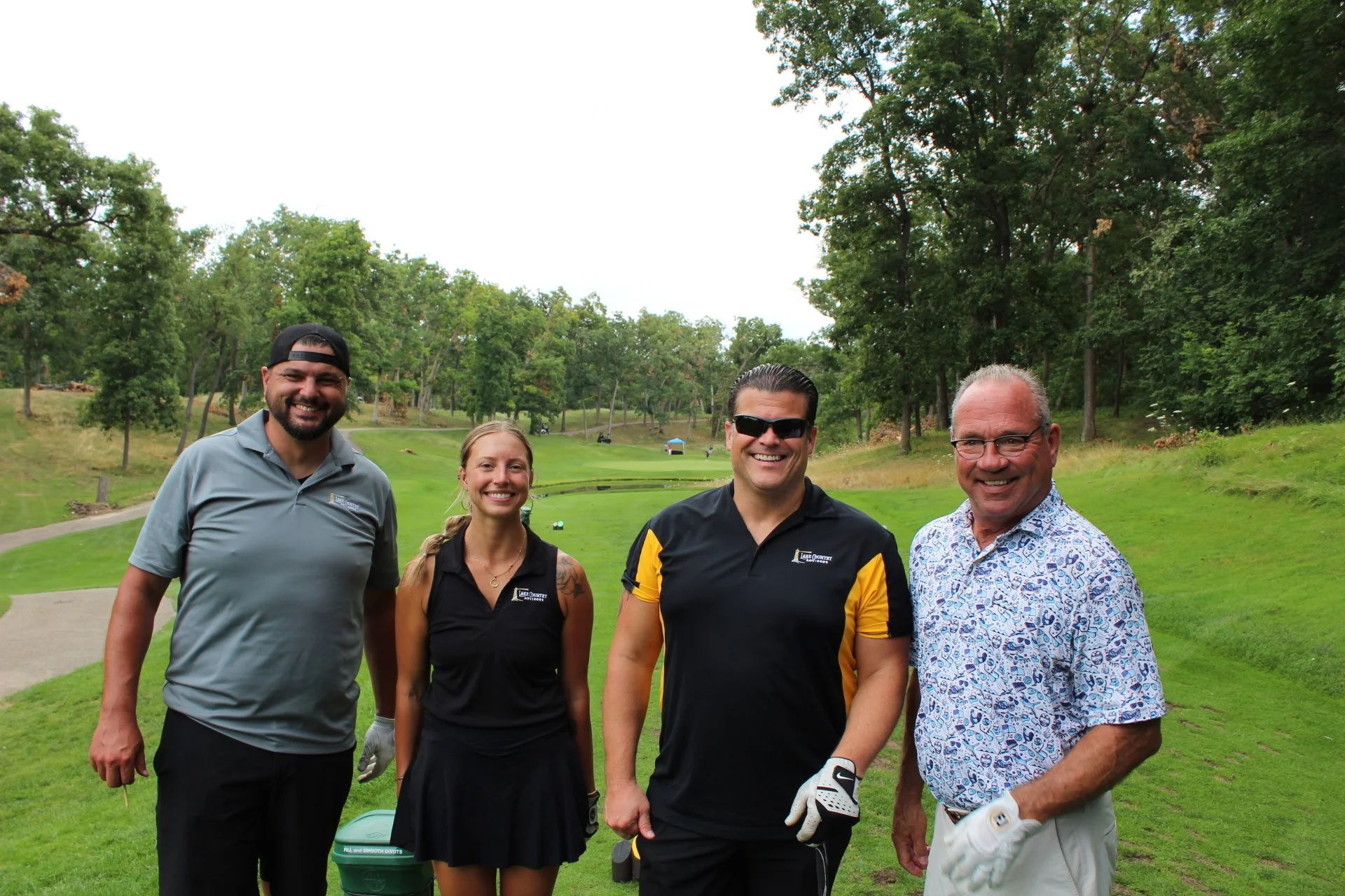 Group of five people, two women and three men, smiling and standing on a golf course with green grass and trees in the background.
