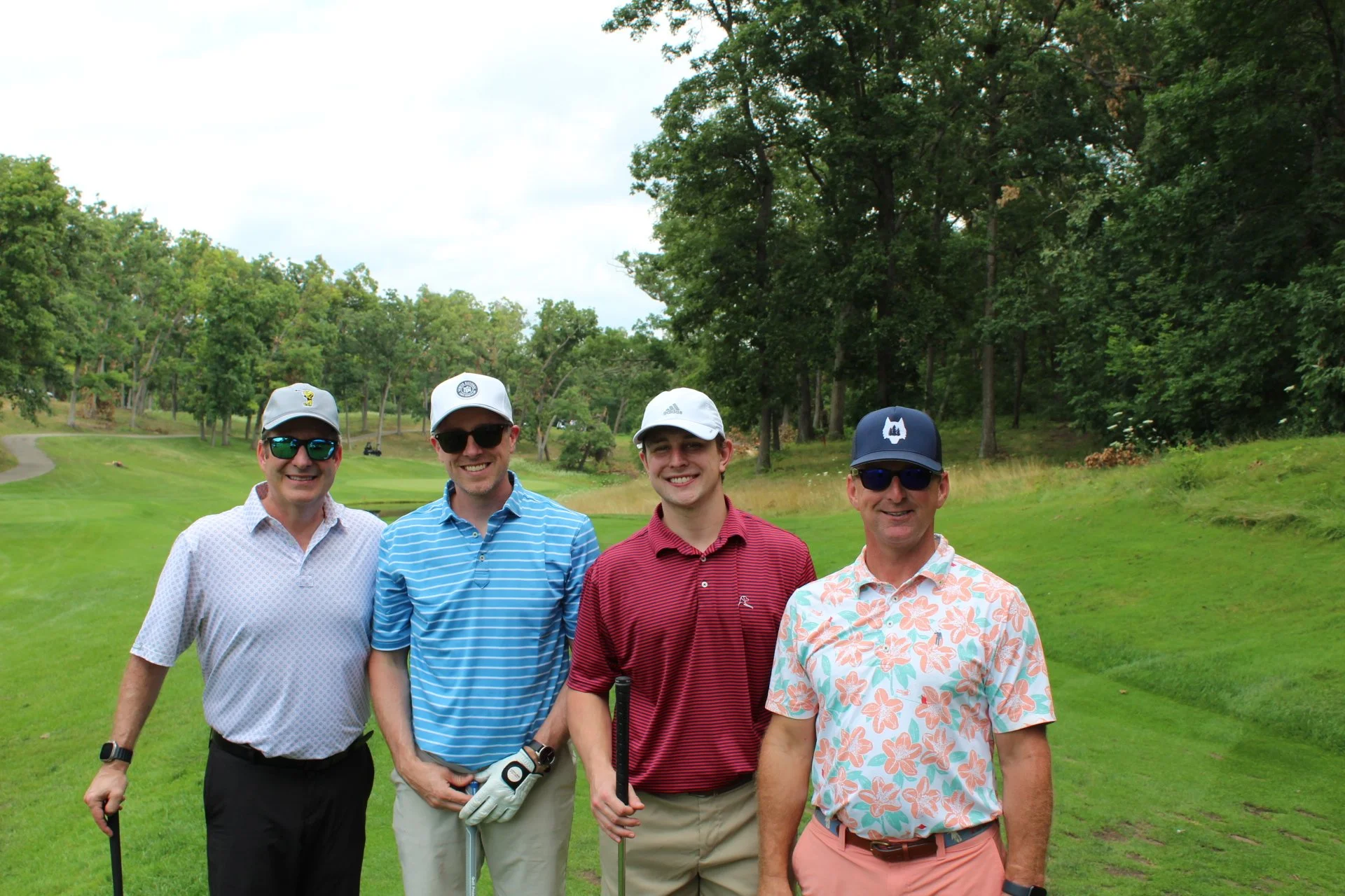 Four men standing on a golf course, smiling, wearing sunglasses and golf caps, with trees and a path in the background.