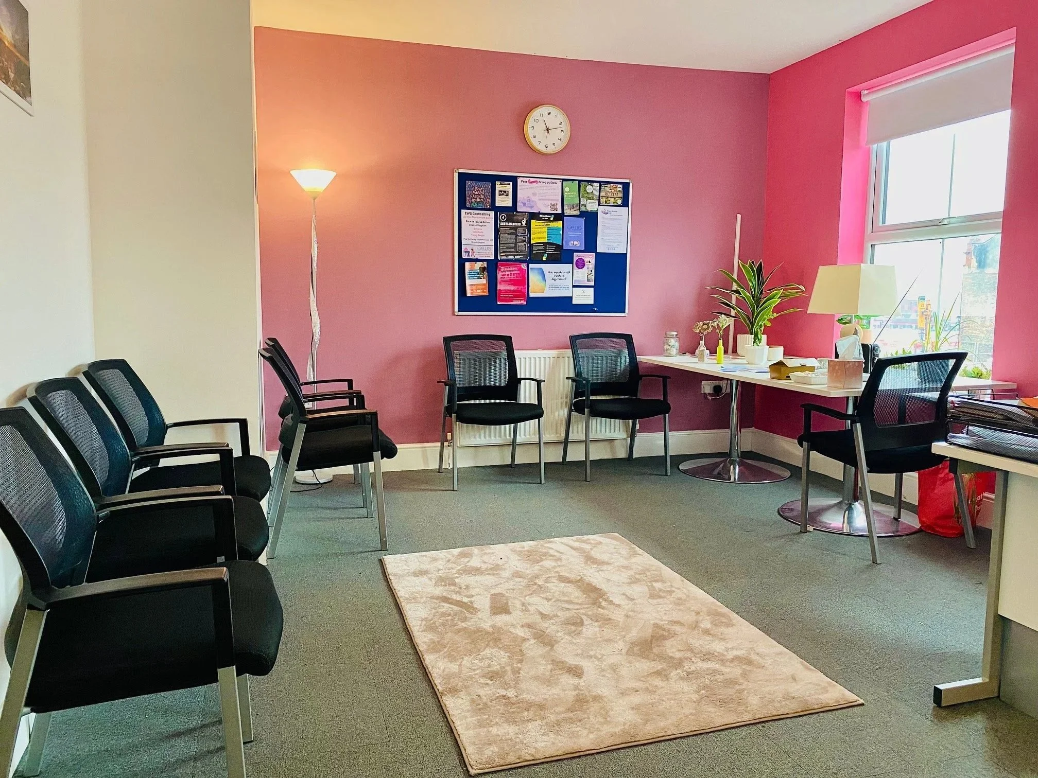 Empty waiting room with six black chairs, pink and white walls, a beige rug, a white desk with a lamp, plants, and a cork bulletin board.