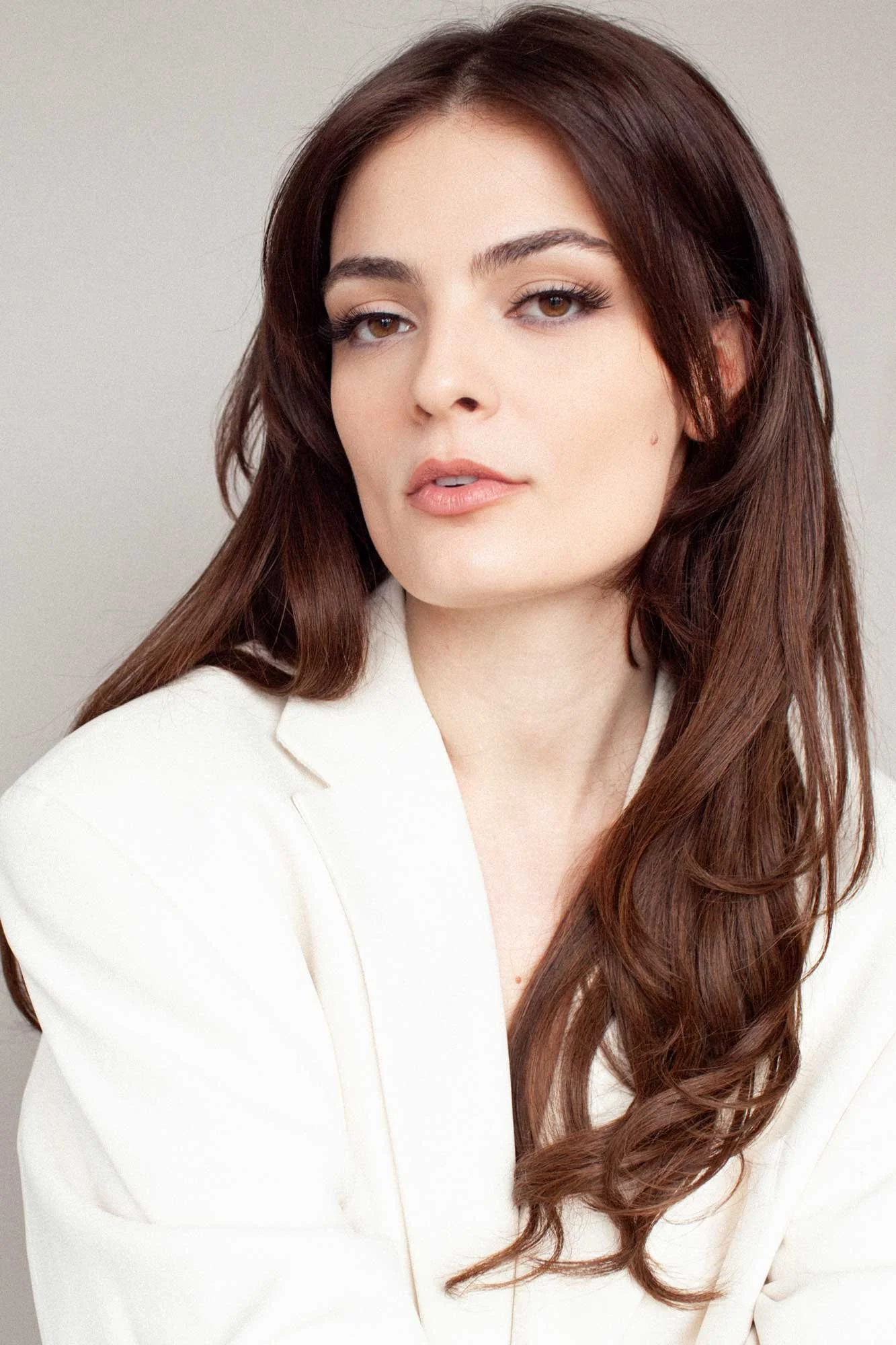 A professional headshot for an actress, director and producer, showing a young woman with pronounced jaw line and eyebrows, long wavy dark hair, wearing a white blazer with big shoulders, photographed in a Brooklyn portrait studio with natural light 