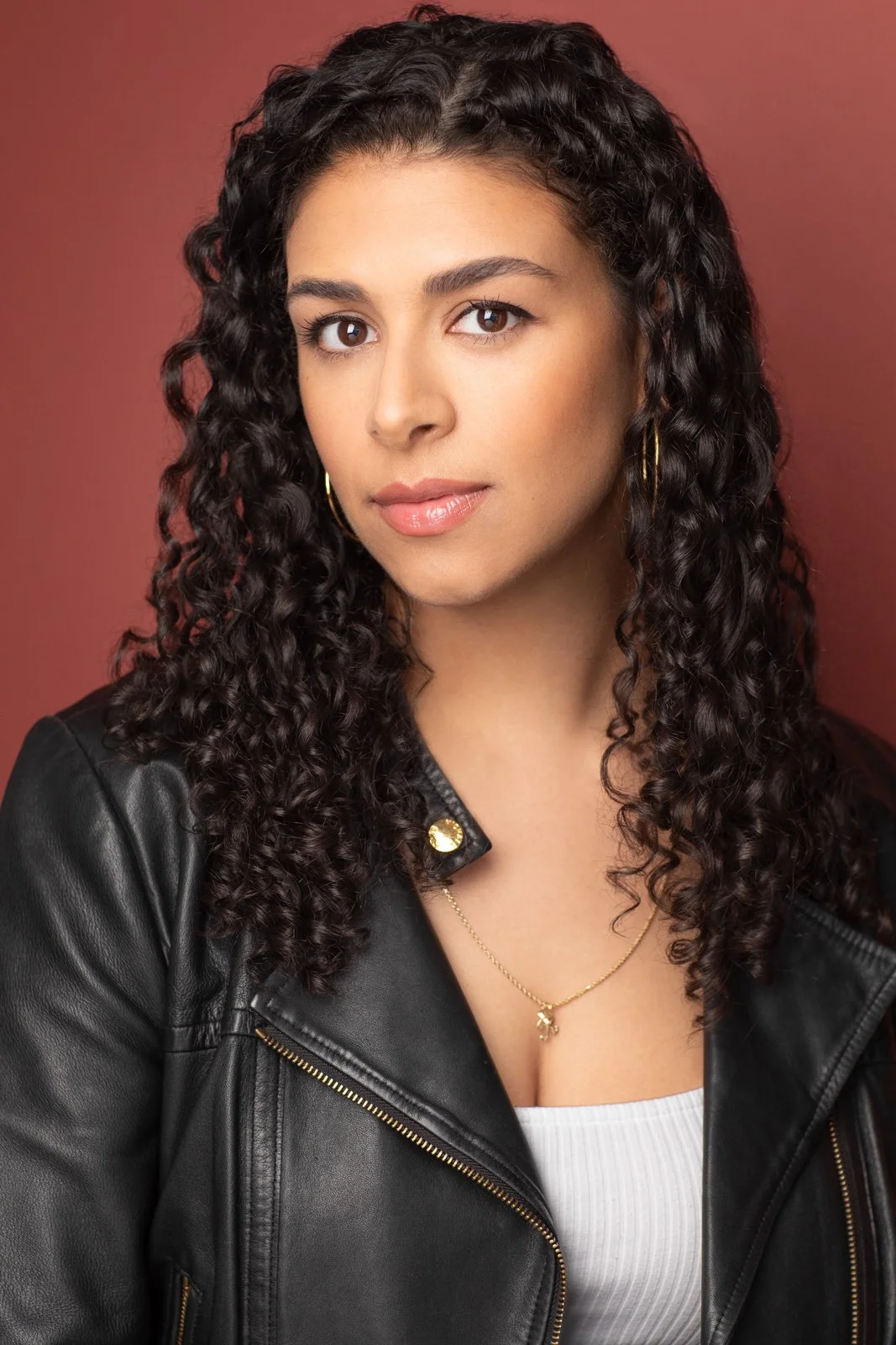 Actor headshot by Hancock Headshots in NYC. A woman with curly dark hair, wearing a black leather jacket, white top, gold necklace and earrings, posing against a red background.