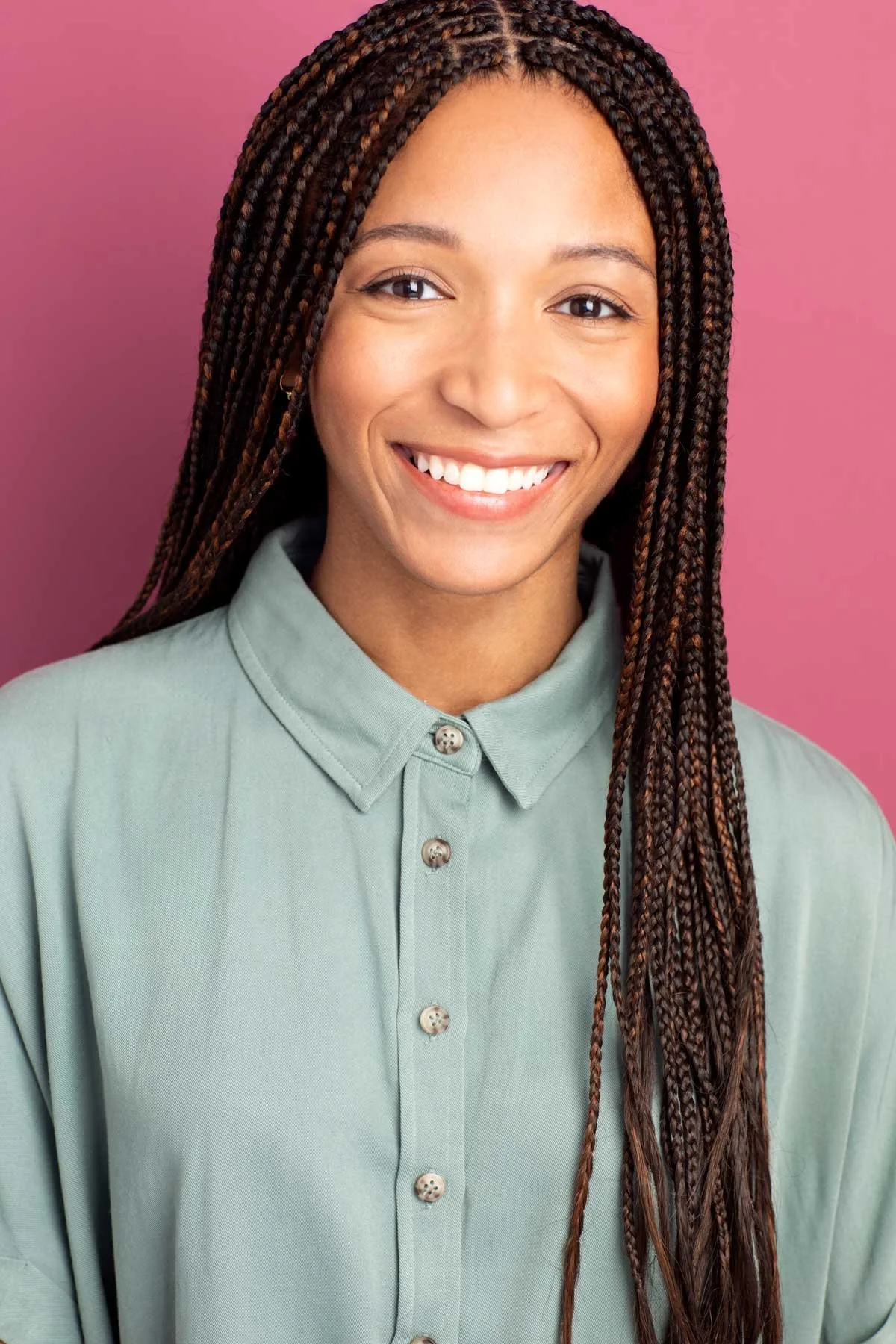 A colorful commercial headshot of a young African American Black actress smiling with bright white teeth, long braided hair, wearing a light green button down collared shirt, standing against a pink background, photographed in a Brooklyn photo studio