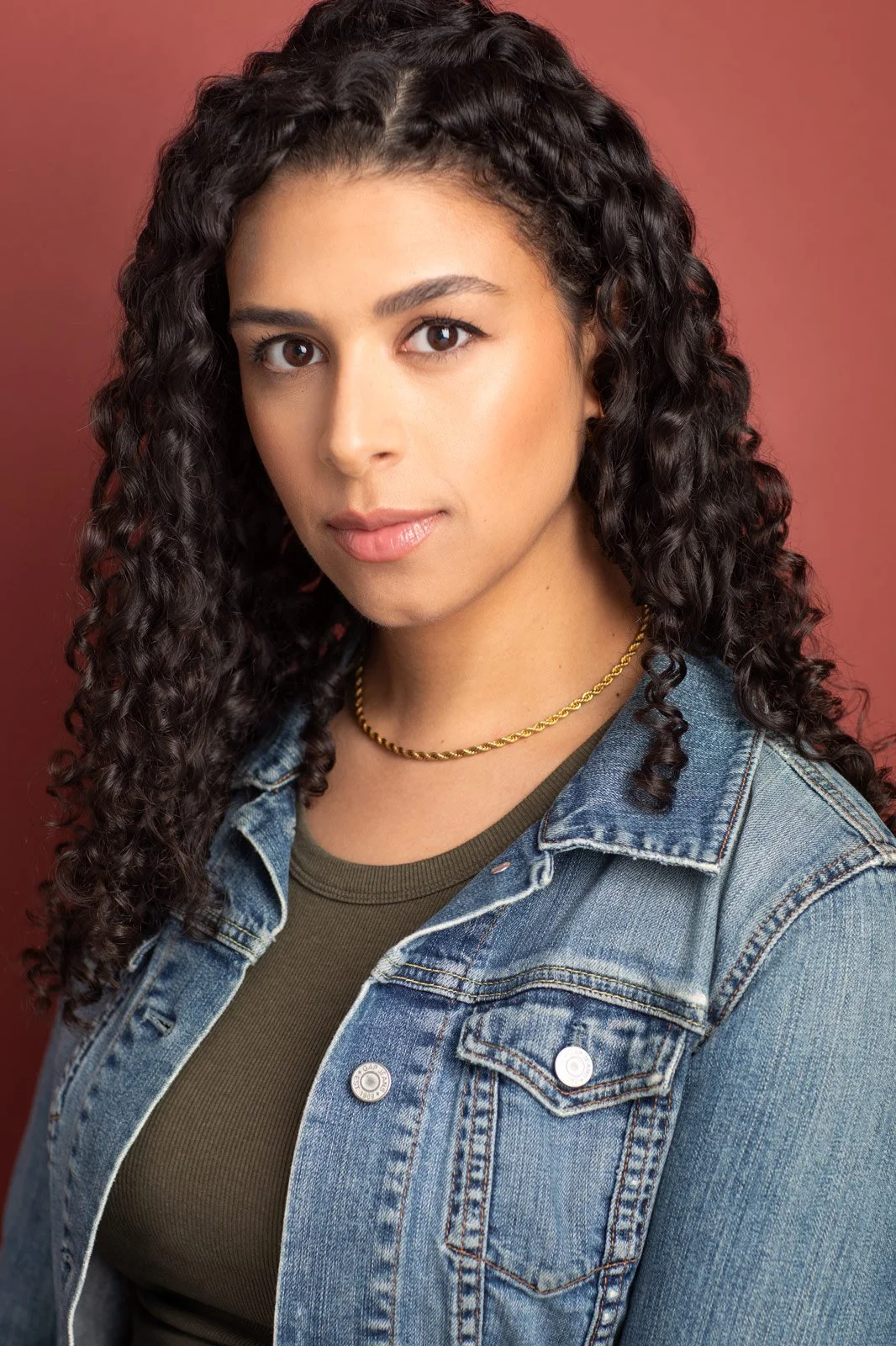 Actor headshot by Hancock Headshots in NYC. A woman with long, curly dark hair, wearing a denim jacket over an olive green top and a gold necklace, poses against a solid reddish background.