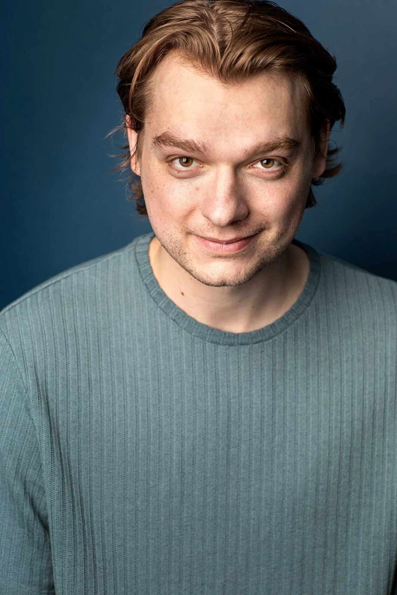 Professional male actor headshot of young man with blonde hair in a sage green sweater with a mischievous smile, natural studio lighting, Brooklyn NYC headshot photographer.