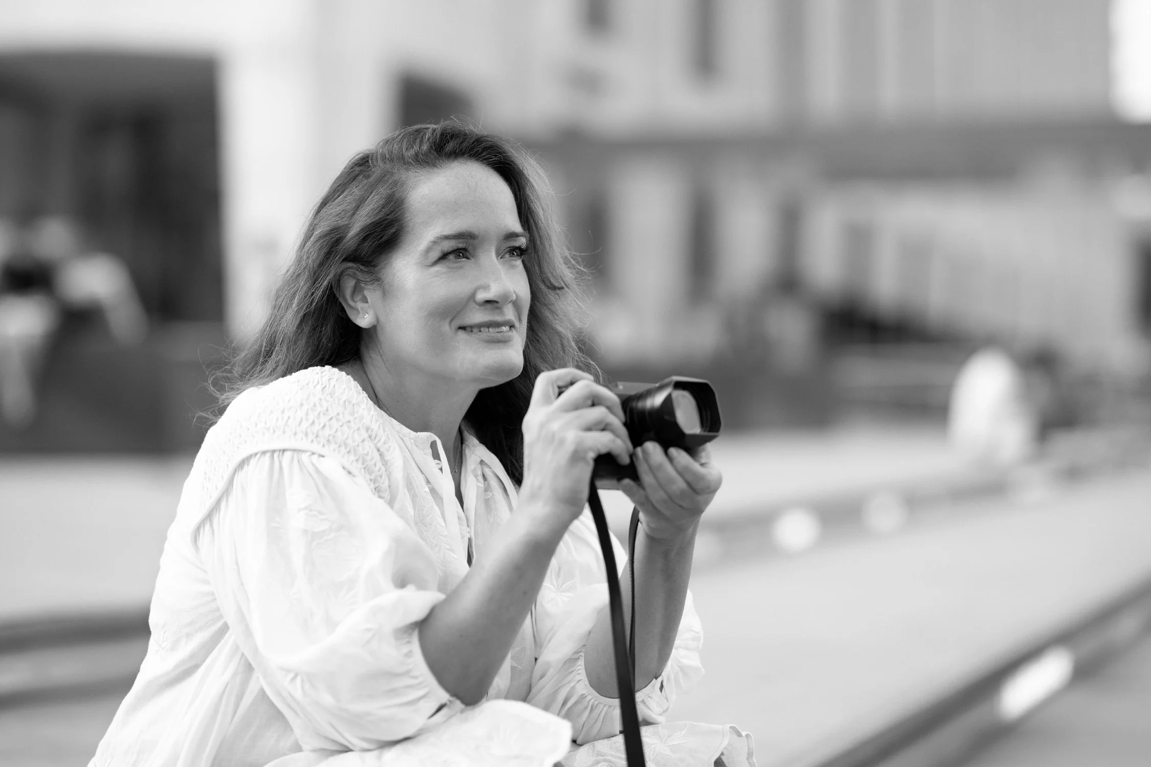 Photographer Lisa Hancock, a woman with long hair in a white blouse holding a camera outdoors, with blurred buildings in the background.