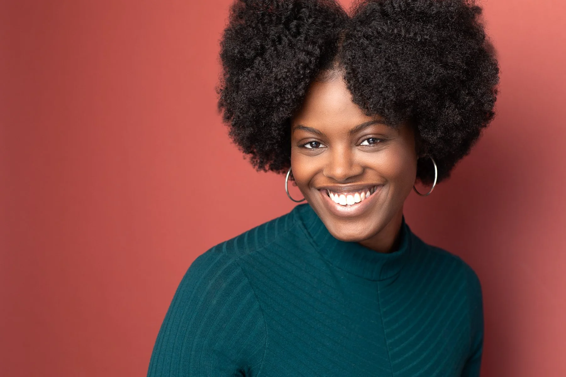 Actor headshot of African American woman photographed by Hancock Headshots.  A smiling woman with natural curly hair, wearing hoop earrings and a teal ribbed turtleneck, against a pink background.