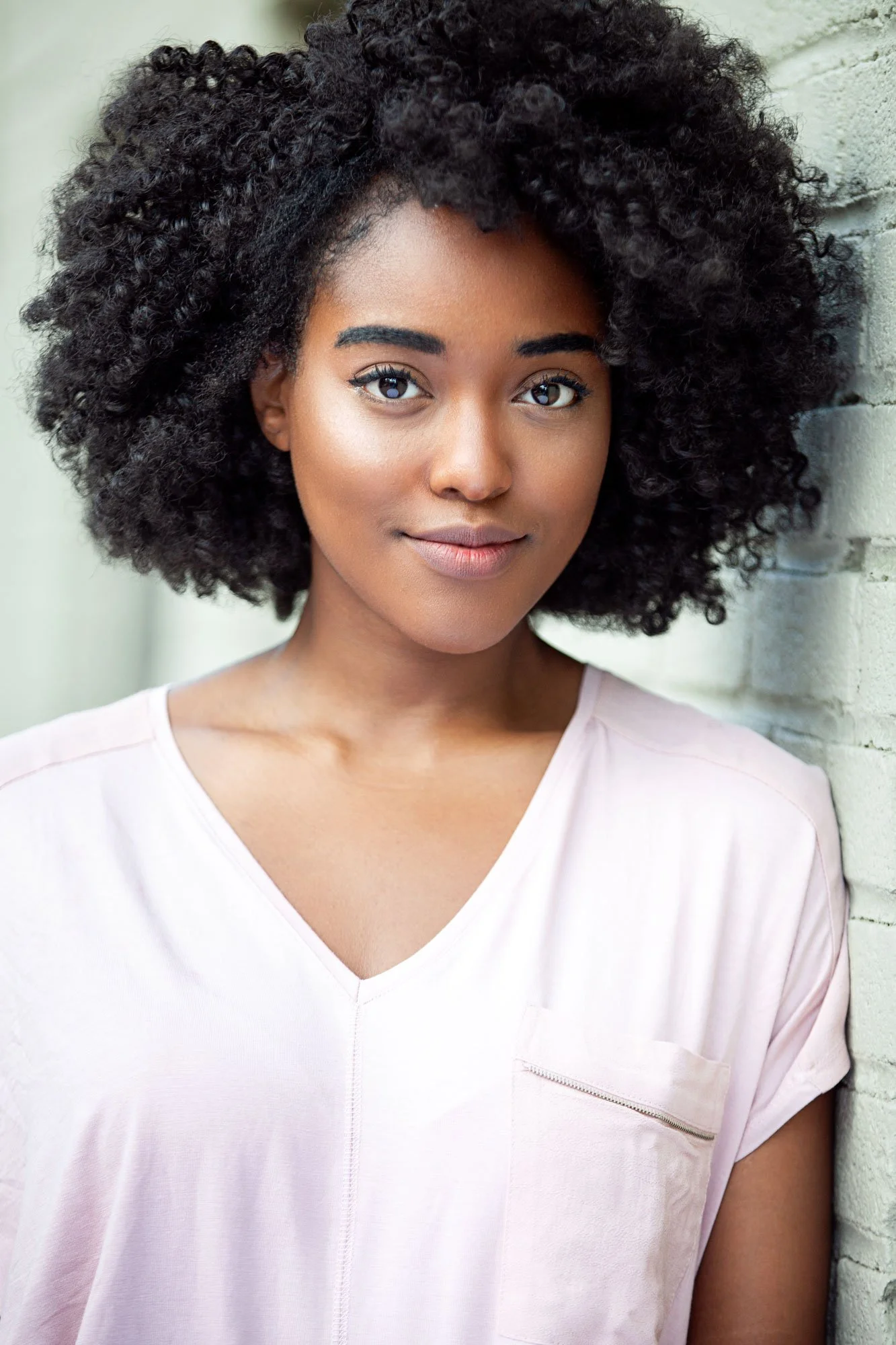 Actor headshot of an African American actress named Arsema Thomas photographed in New York City by Hancock Headshots. A close-up of a woman with dark, curly hair wearing a light pink shirt, standing next to a white brick wall.