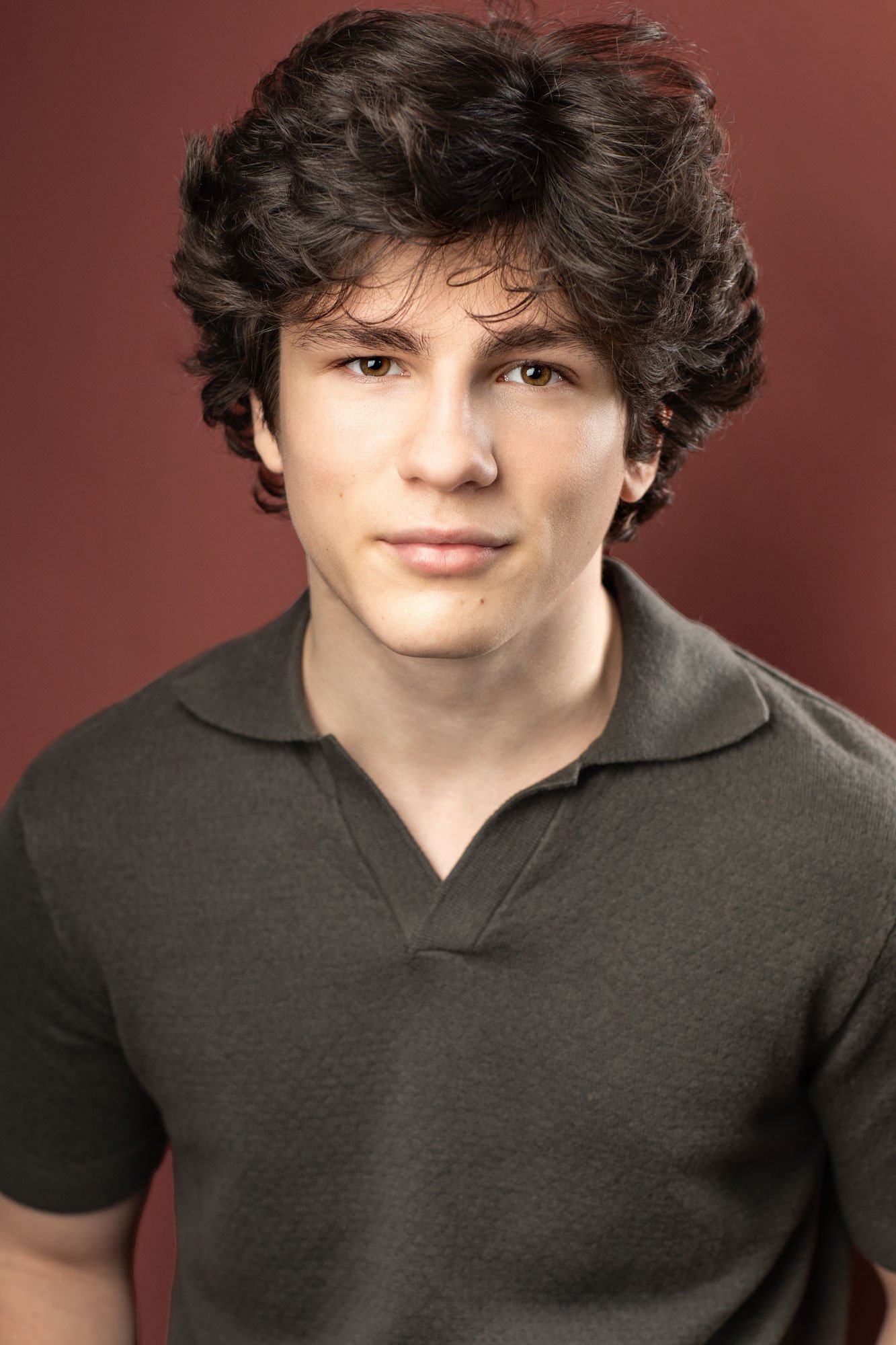 Young male actor headshot photographed in a Brooklyn studio.  A young teenage actor with thick wavy brown hair wears a brown polo-style sweater and a subtle smile, against a dark rust background.