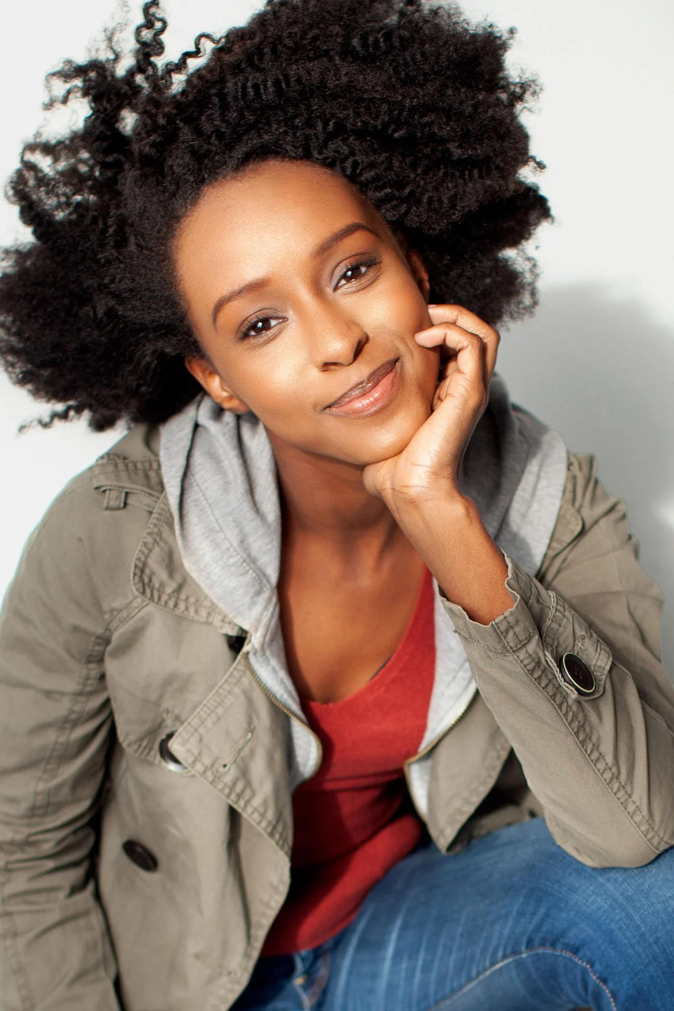 Acting headshot of Ebony Obsidian photographed by Hancock Headshots in Brooklyn, with her chin in her hand, smiling warmly, wearing an army jacket against a white backdrop, her hair natural
