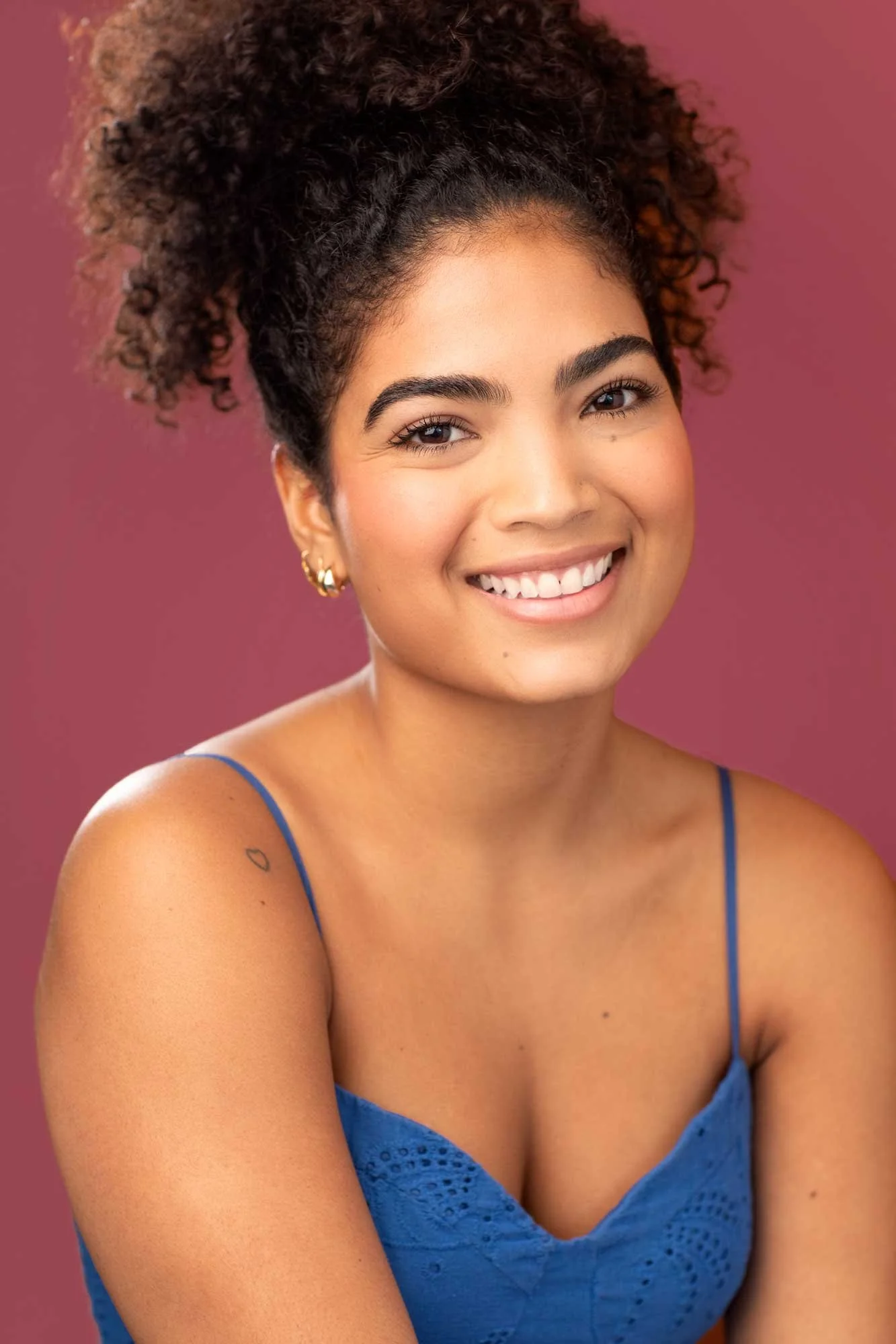 NYC commercial acting headshot of young Latina woman in her early 20s with curly dark hair in a high pony tail, smiling broadly, wearing a blue sundress photographed against a pink background