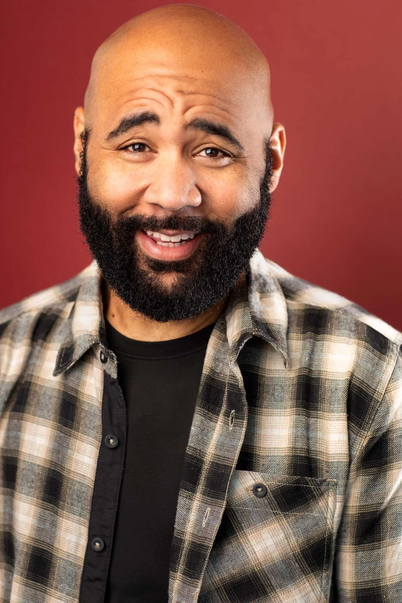 Bearded black actor in plaid flannel and black tee laughing warmly against dark red backdrop — Brooklyn rom-com and comedy headshot photographed at Hancock Headshots.