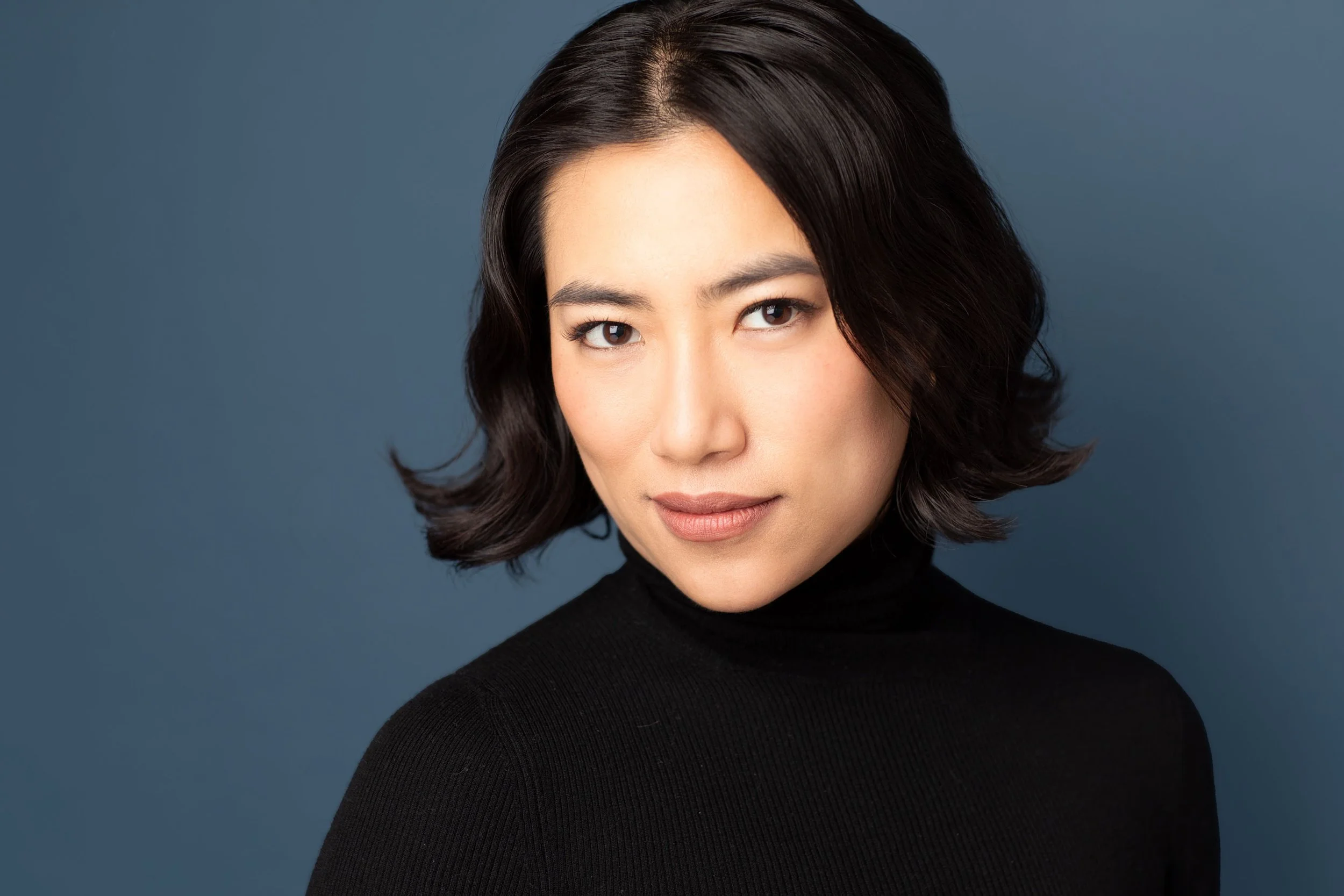 Legit actor headshot photographed in NYC by Hancock Headshots, Asian woman with bob against dark blue backdrop wearing black turtle neck