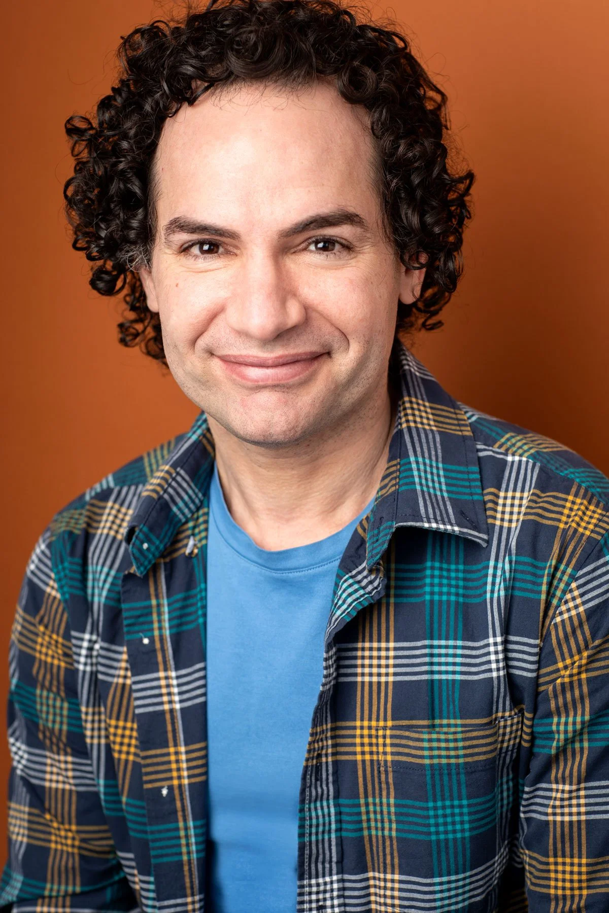 Actor headshot photographed by Hancock Headshots in NYC, commercial shot of man with curly dark hair smiling against a vibrant orange backdrop