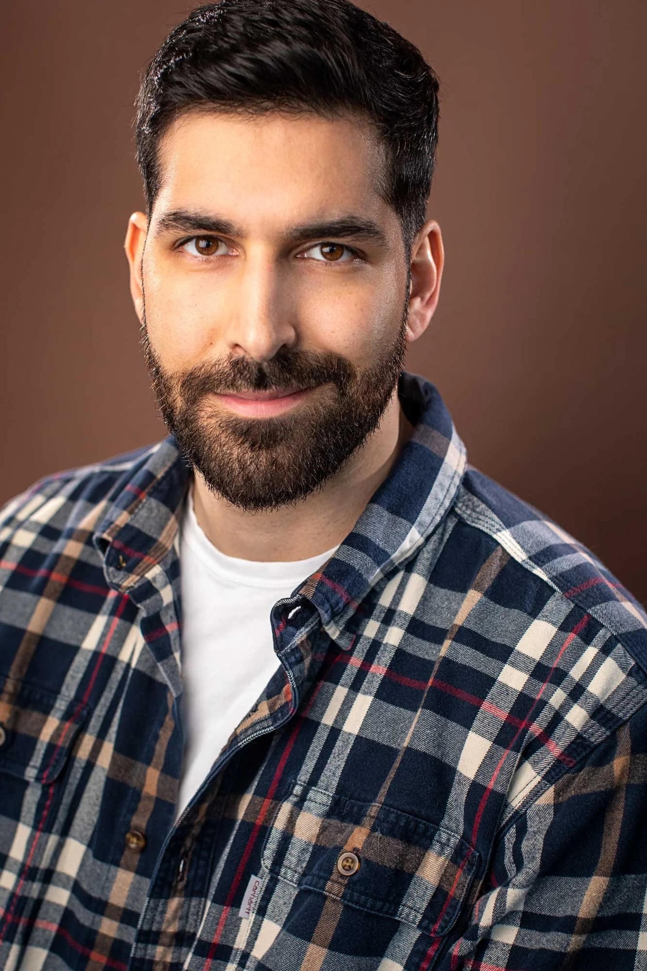 Acting headshot of young man with dark brown hair, moustache and beard, wearing a blue flannel shirt, photographed in Brooklyn against a brown background