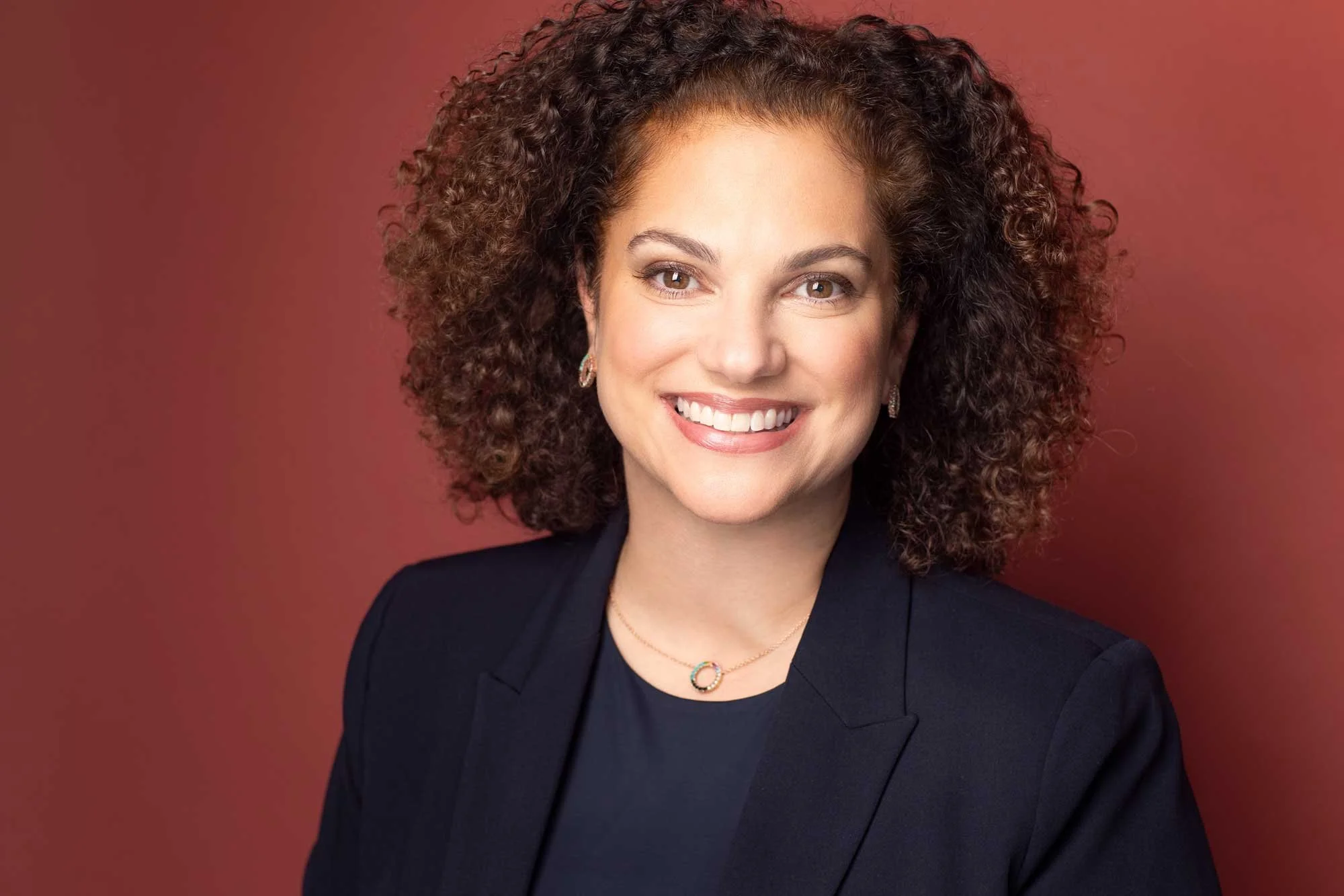 Landscape-oriented corporate headshot of a woman with curly brown hair wearing a black blazer and a necklace, smiling against a rust-colored backdrop, photographed in a Brooklyn headshot studio.
