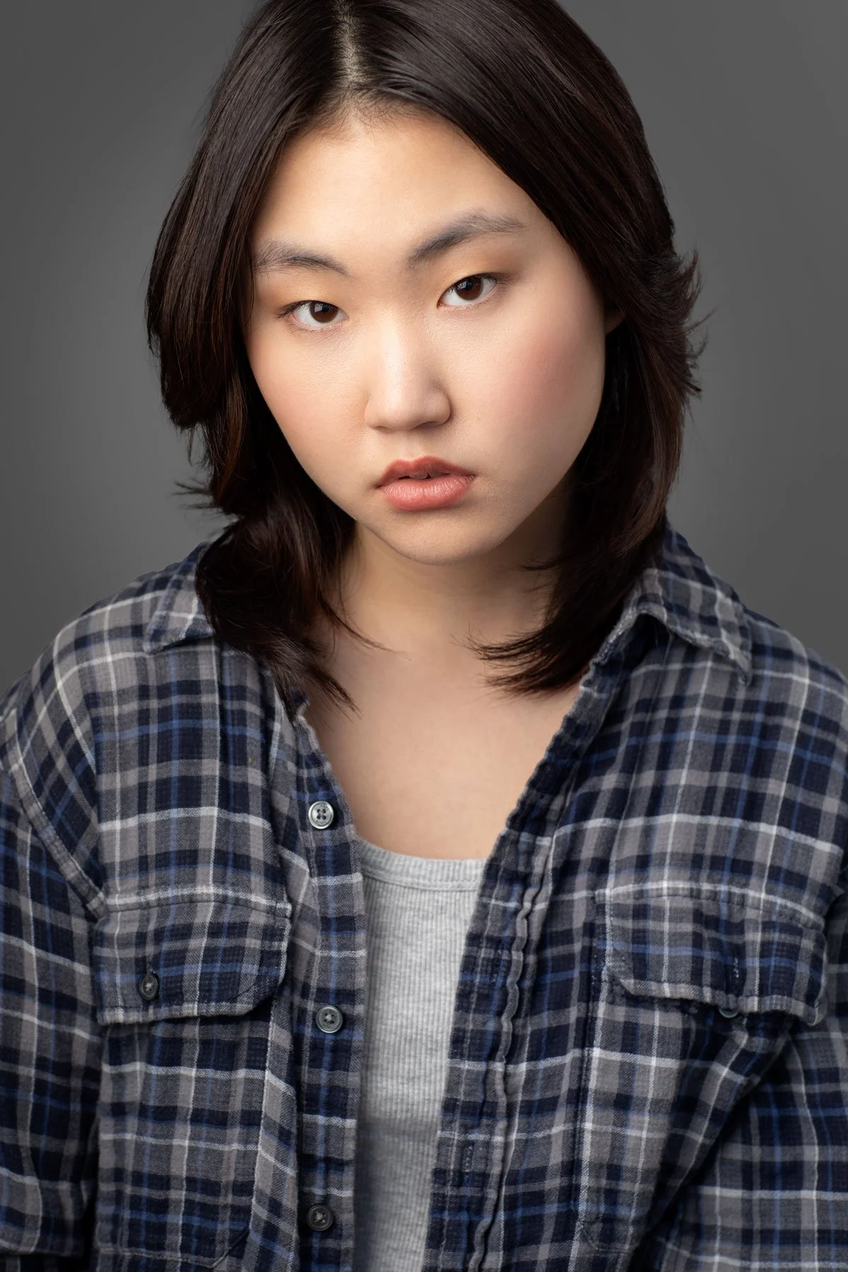 Portrait of a young woman with dark hair wearing a plaid shirt and a white top against a gray background.