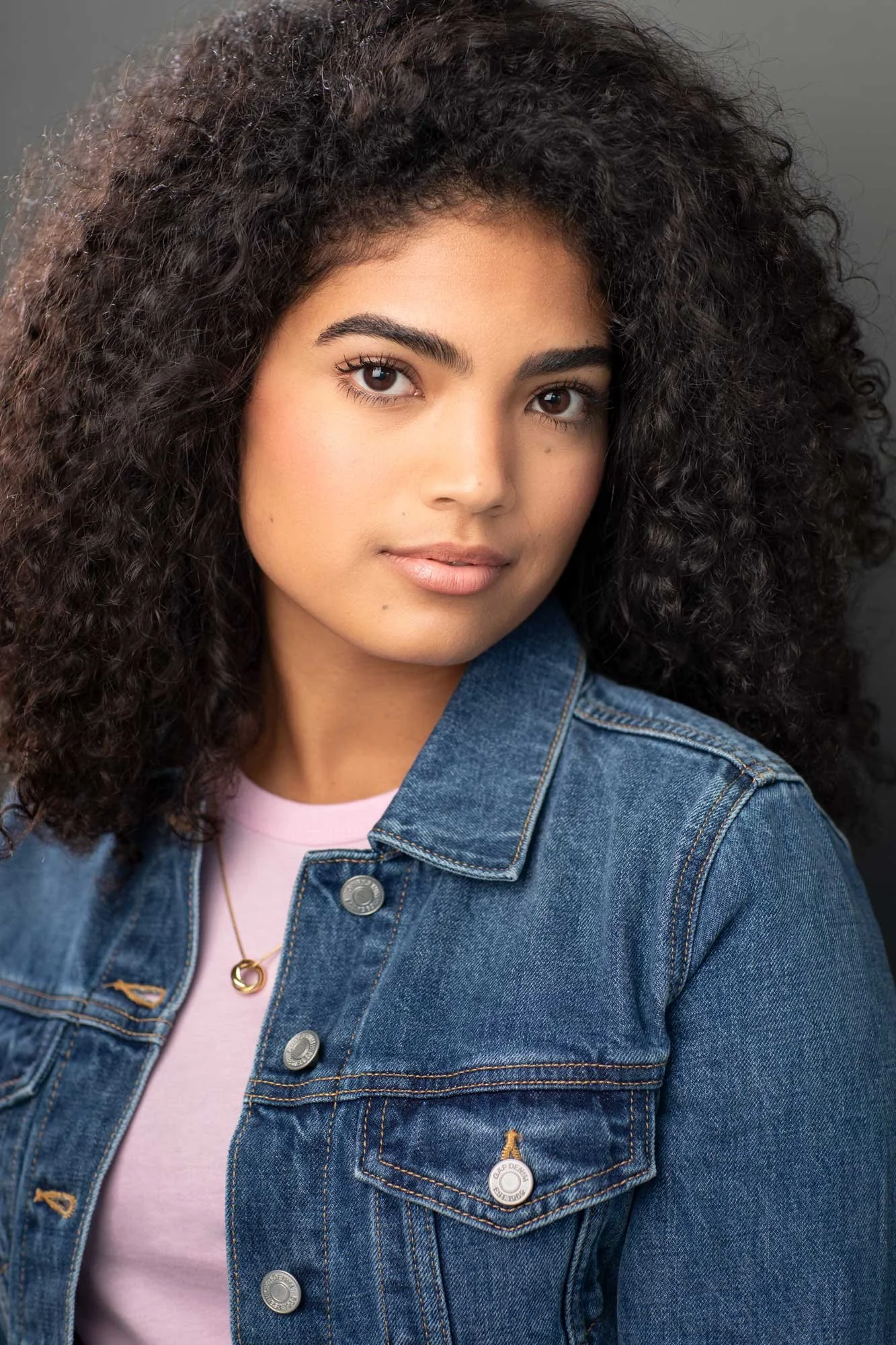 Dramatic acting headshot of a young Latina actress, wearing a denim jacket over a pink t-shirt, with natural curly hair, photographed against a dark grey background in a Brooklyn studio.