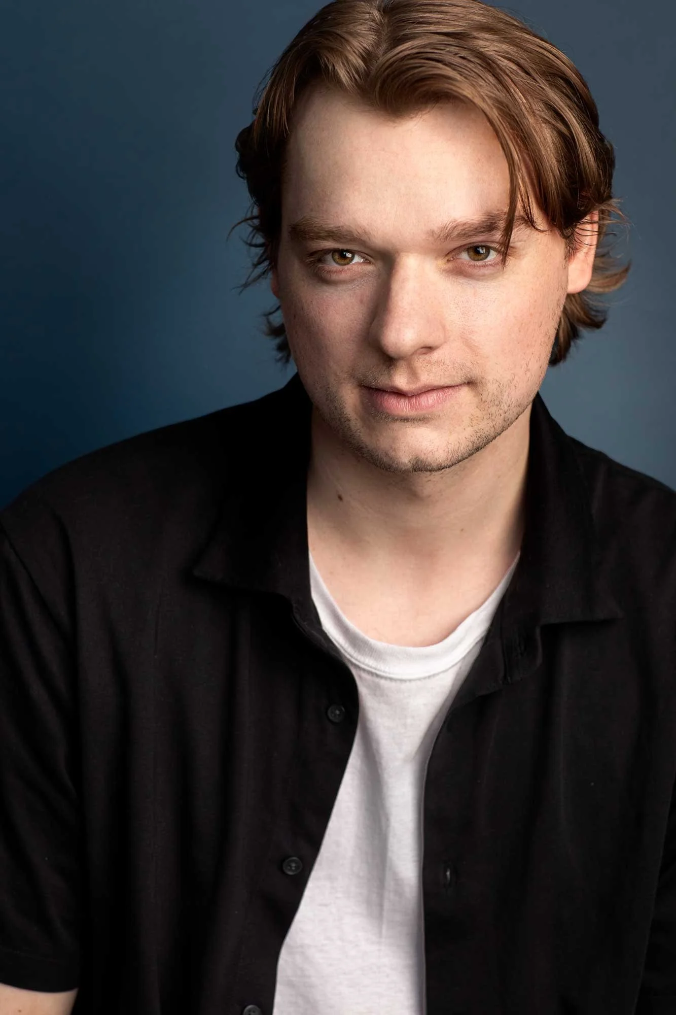 Charismatic male actor headshot in black shirt over white t-shirt, smoldering confident expression, romantic lead casting photo, NYC.
