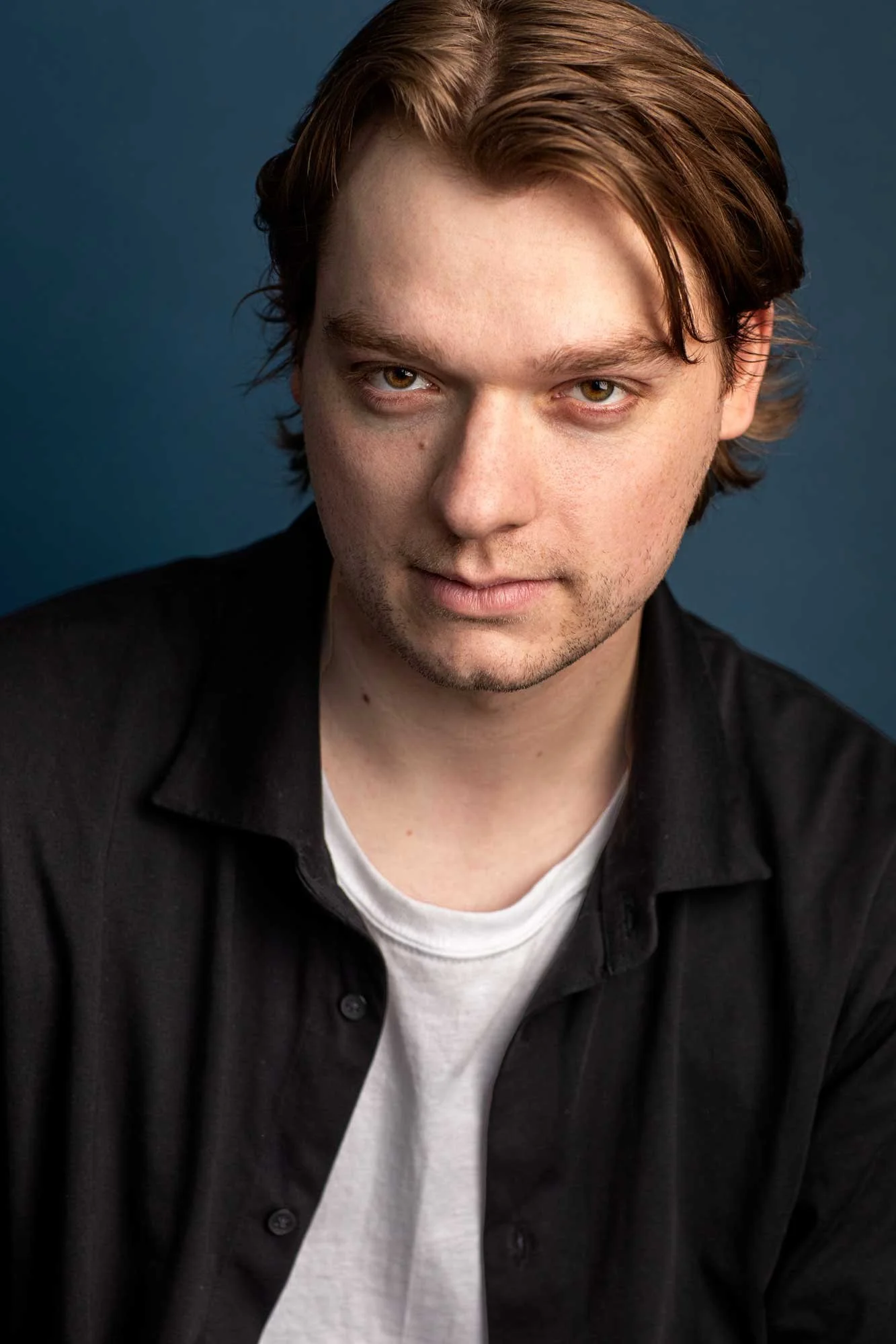 Professional Brooklyn actors headshot showing dramatic young man staring intensely into the camera against a moody dark blue background, wearing a dark jacket over white t-shirt with tousled dirty blonde hair falling in front of his eyes.