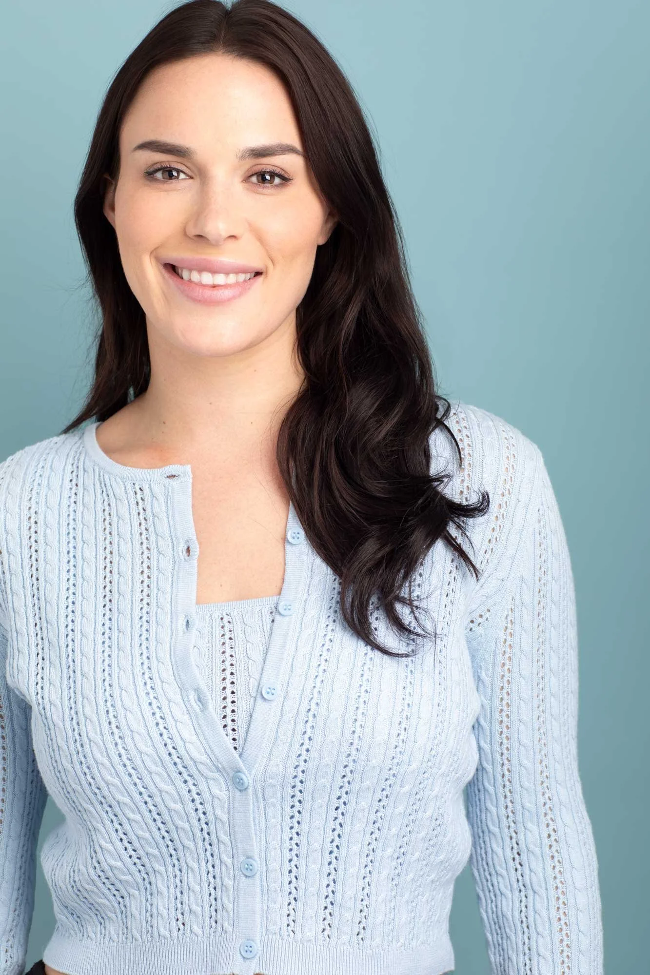 A brightly colored commercial headshot of a smiling actress and model with long dark hair wearing a baby blue cardigan over matching knit top, photographed in a Brooklyn photo studio against a light blue backdrop.