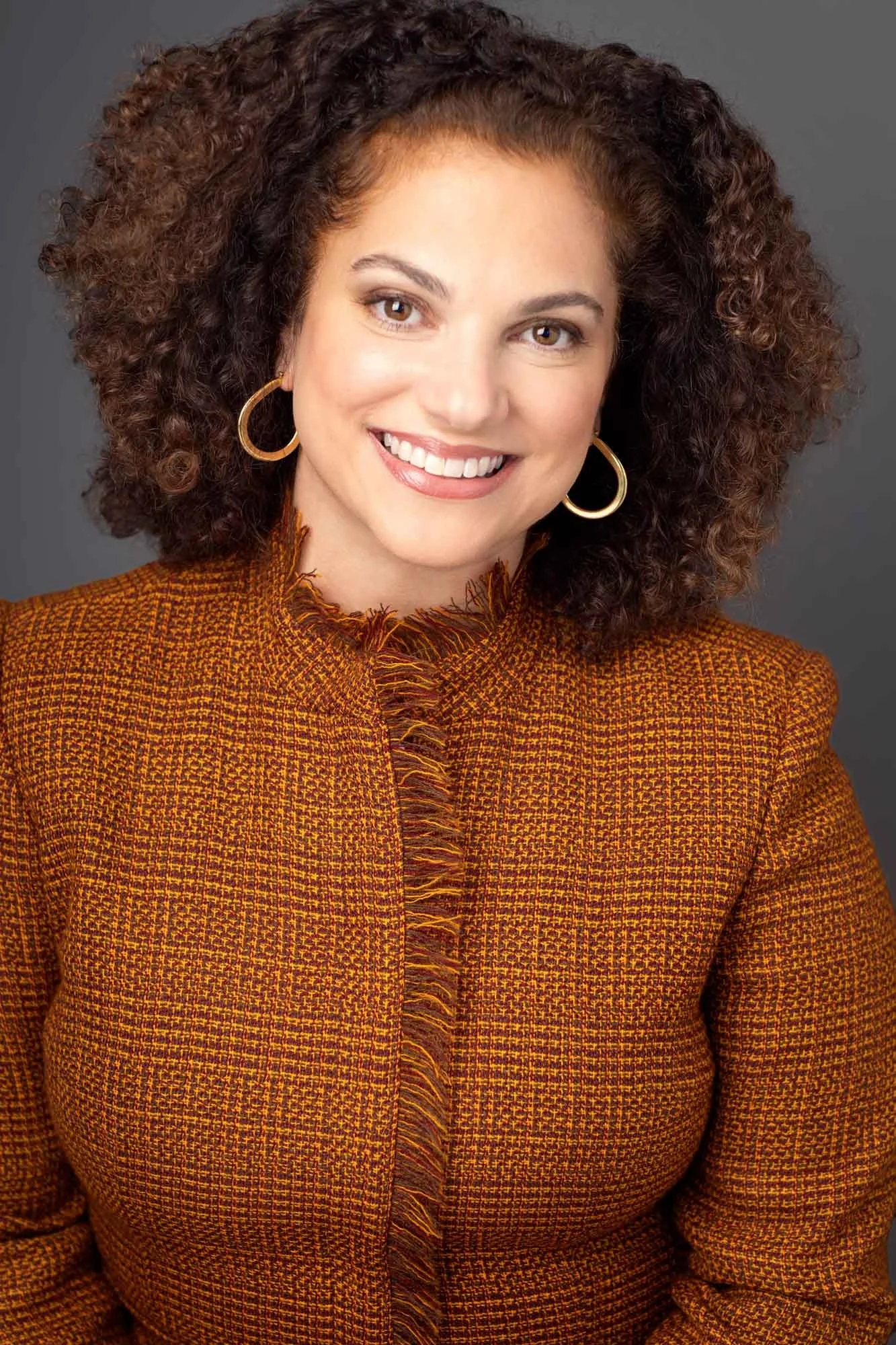 Corporate headshot of a woman in her 40s with brown curly hair and light skin smiling, wearing gold hoop earrings and an orange tartan jacket, photographed in a Brooklyn photography studio against a classic grey backdrop.
