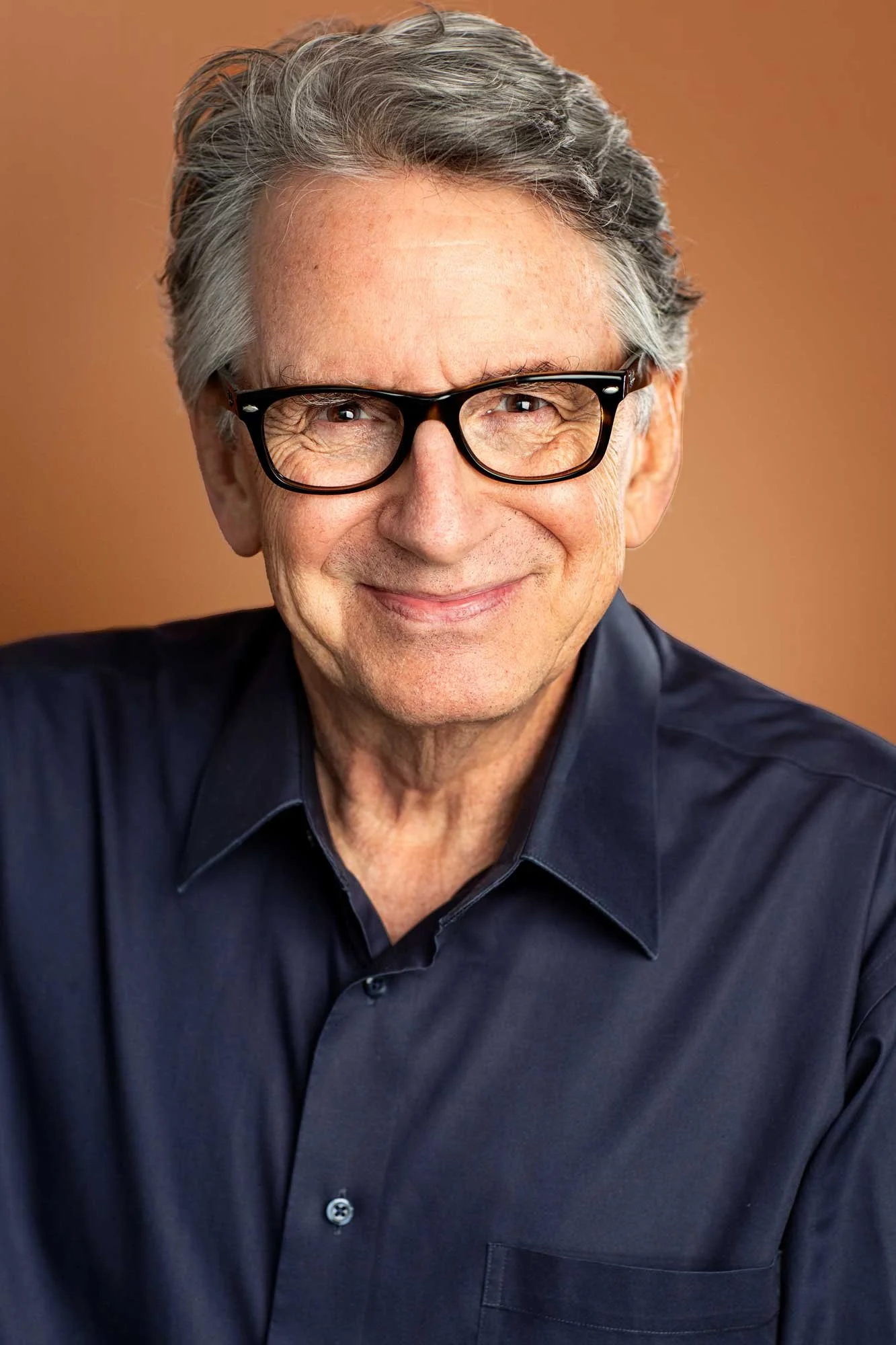 Acting headshot of elderly man in his 60s with dark grey hair wearing a navy button down shirt, dark-framed glasses, smiling radiantly, photographed in a Brooklyn headshot photographer's studio.