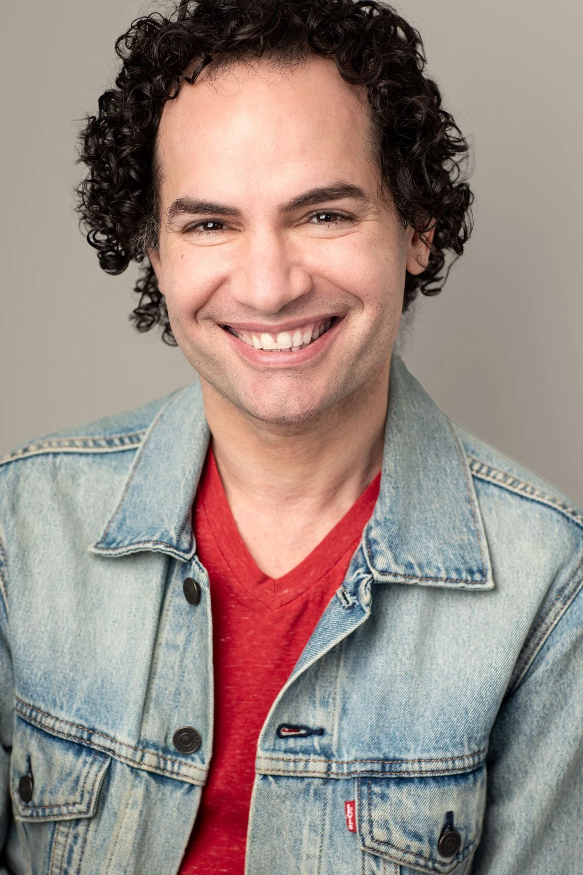 Actor headshot photographed by Hancock Headshots in NYC, commercial shot of man with curly dark hair smiling broadly against a bright neutral backdrop