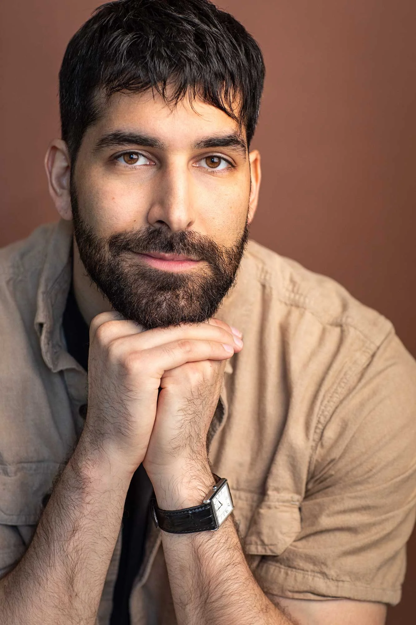 Acting headshot of young man with dark brown hair, moustache and beard, resting his chin on his hands, smirking slightly, wearing a tan shirt, photographed in Brooklyn against a brown background