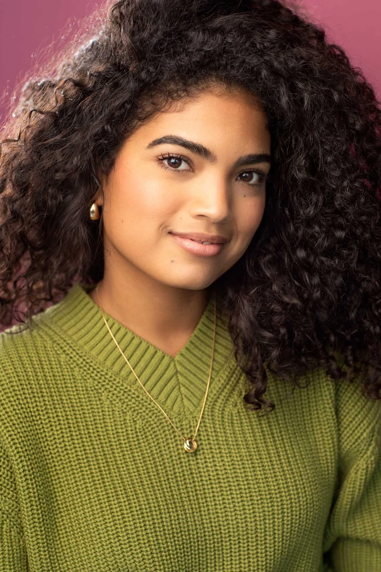 Acting headshot of young Latina actress wearing a green sweater, a big head of natural curly hair, smiling gently, photographed in New York City against a pink background.