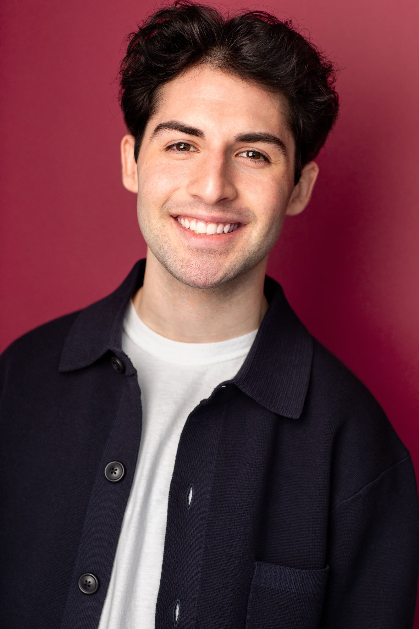 A young man with dark, voluminous hair and a bright smile, wearing a white shirt and a dark jacket, stands against a solid pink background.