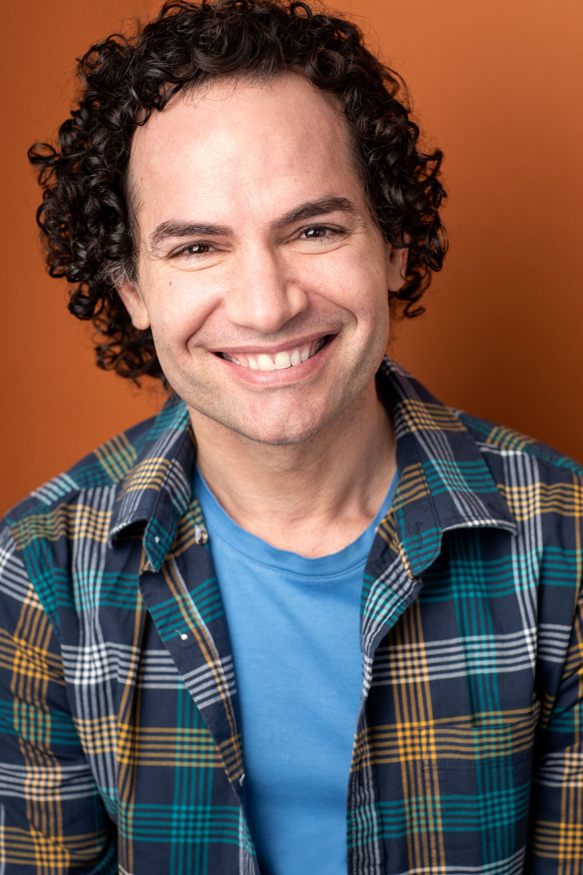 Actor headshot photographed by Hancock Headshots in NYC, commercial shot of man with curly dark hair smiling broadly against a vibrant orange backdrop