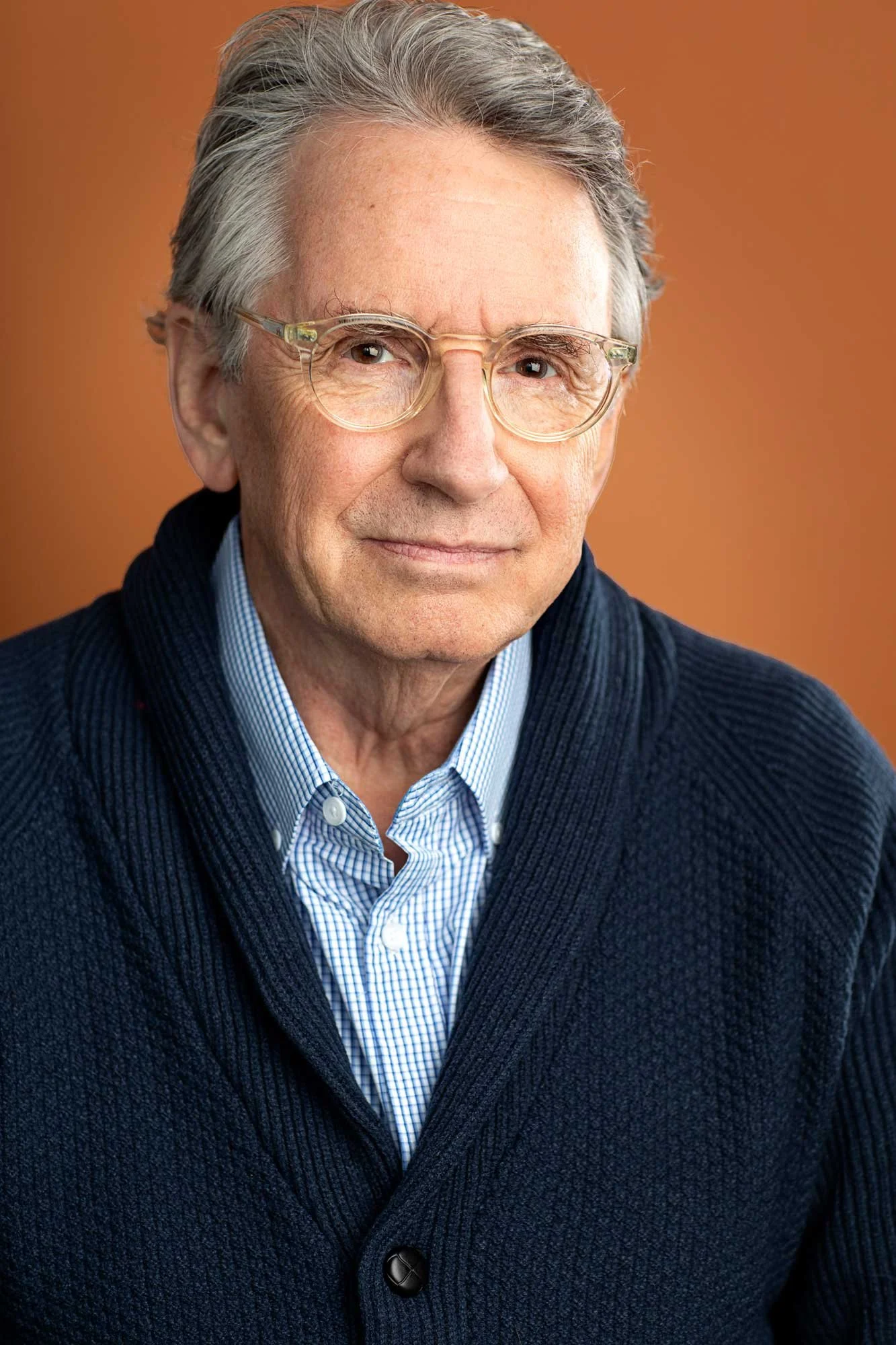 Acting headshot of a senior actor, an older distinguished man wearing clear acetate frames, a shawl collar blue cardigan, and gingham button down underneath — photographed against an orange background in a Brooklyn studio. 