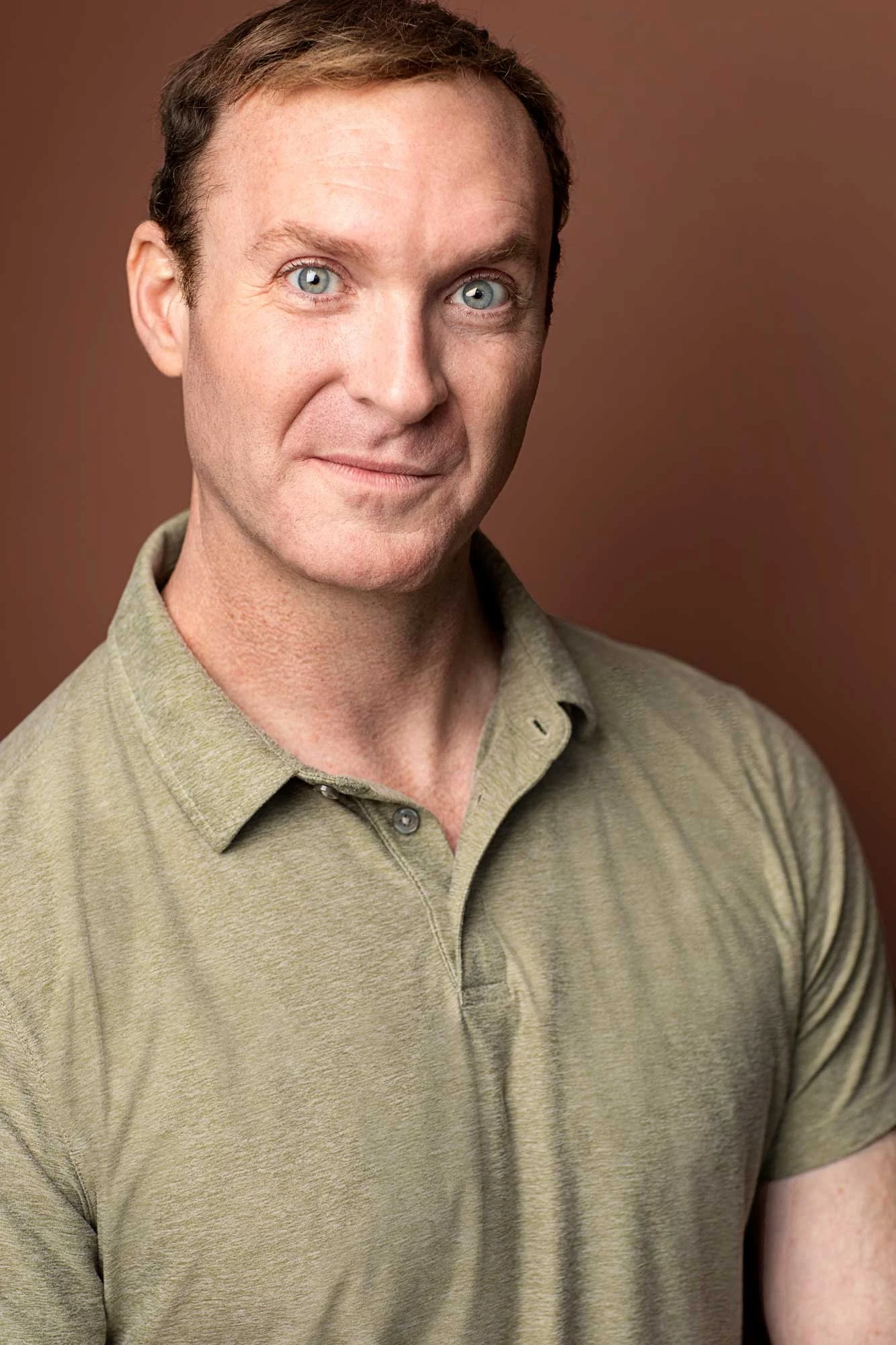 A man with short brown hair, a bright twinkle in his blue eyes, and fair skin is photographed for an actor headshot in a Brooklyn studio, wearing a light olive green polo shirt against a plain brown background.
