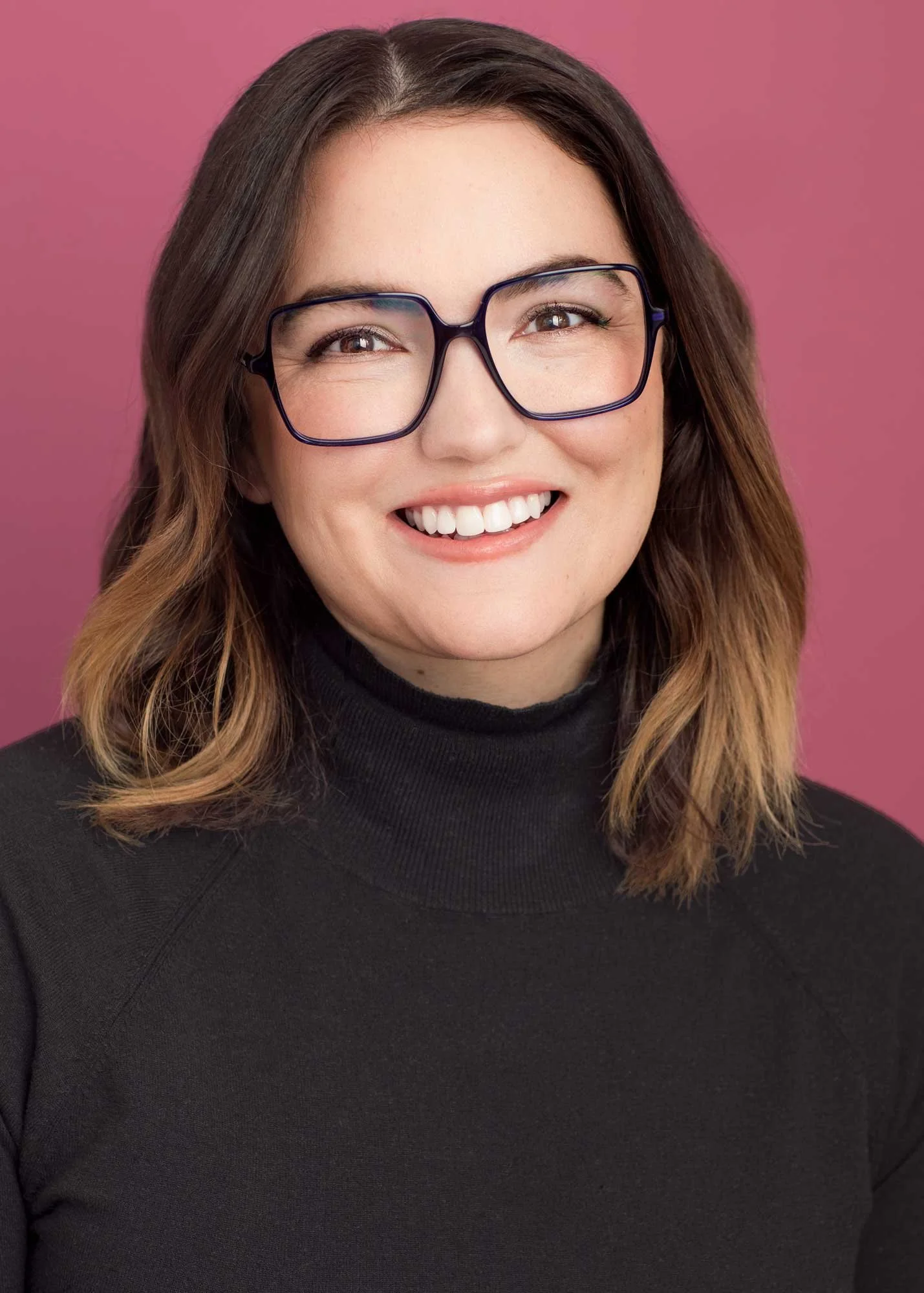 Unconventional business headshot for LinkedIn of a smiling woman with wavy brown hair, wearing dark glasses and a black turtleneck, against a pink background, photographed in Brooklyn studio.
