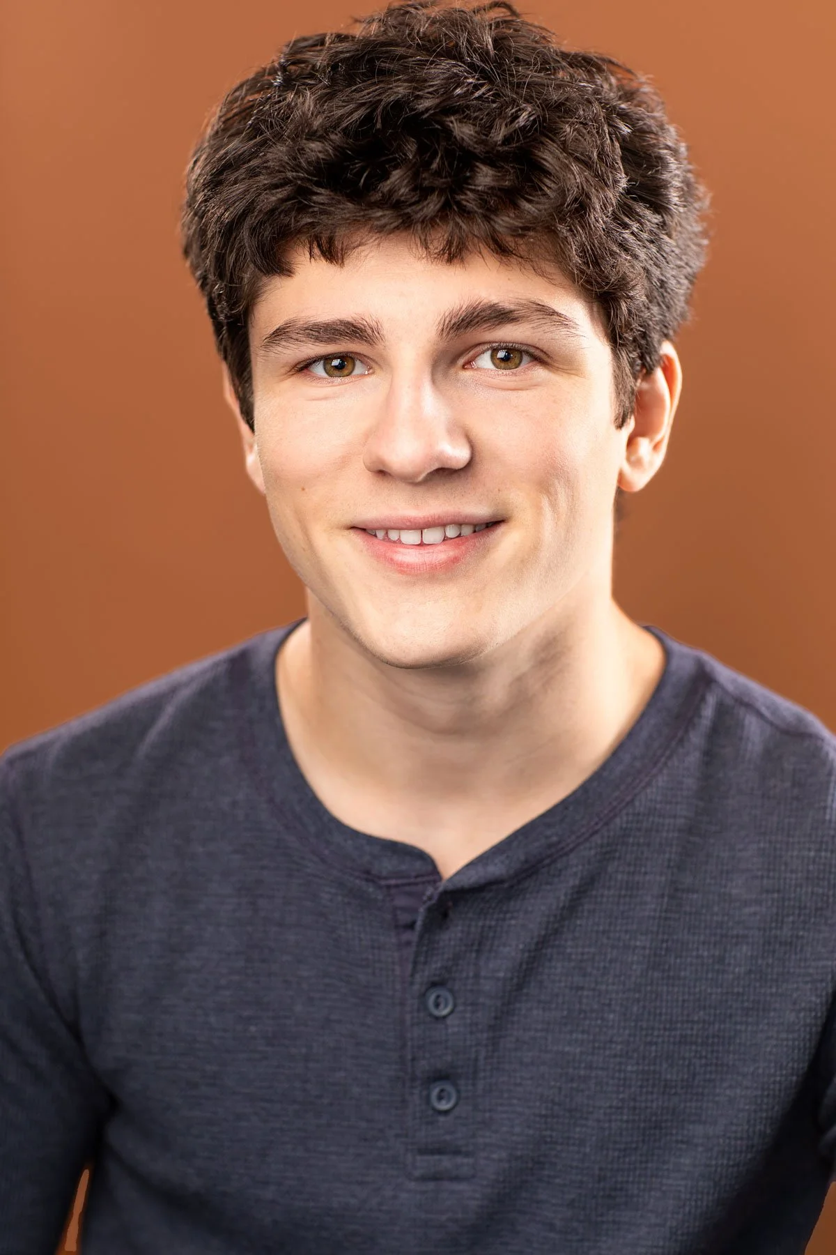 NYC commercial acting headshot of young male actor with a warm, genuine smile, wearing a navy henley against a brown backdrop photographed at Hancock Headshots in Brooklyn