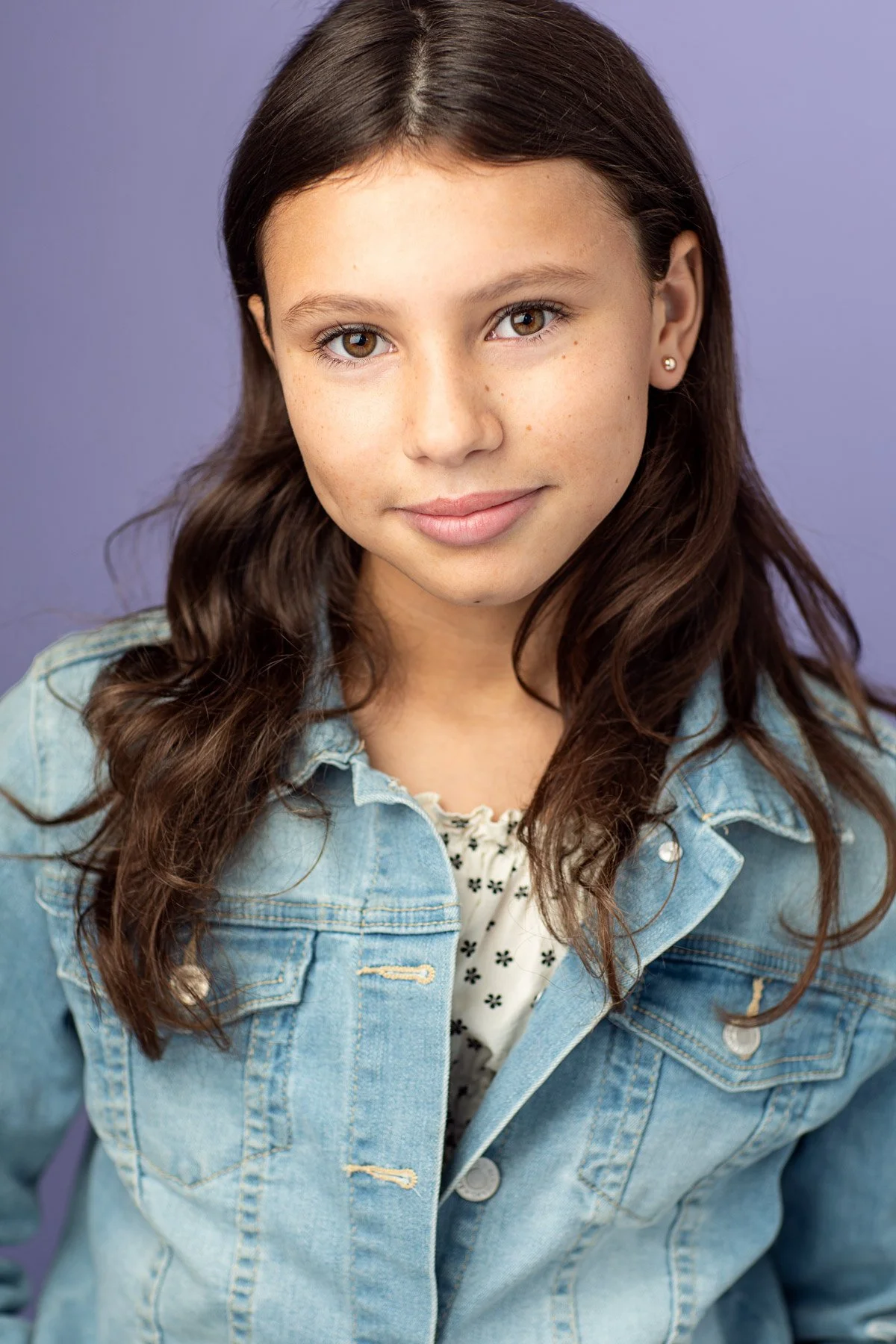 Professional acting headshot of a young girl about 12 years old, smiling gently into the camera, with long brown hair tucked behind her ears, wearing a light blue jean jacket over a ruffled white top with tiny black flowers, photographed in a Brookly