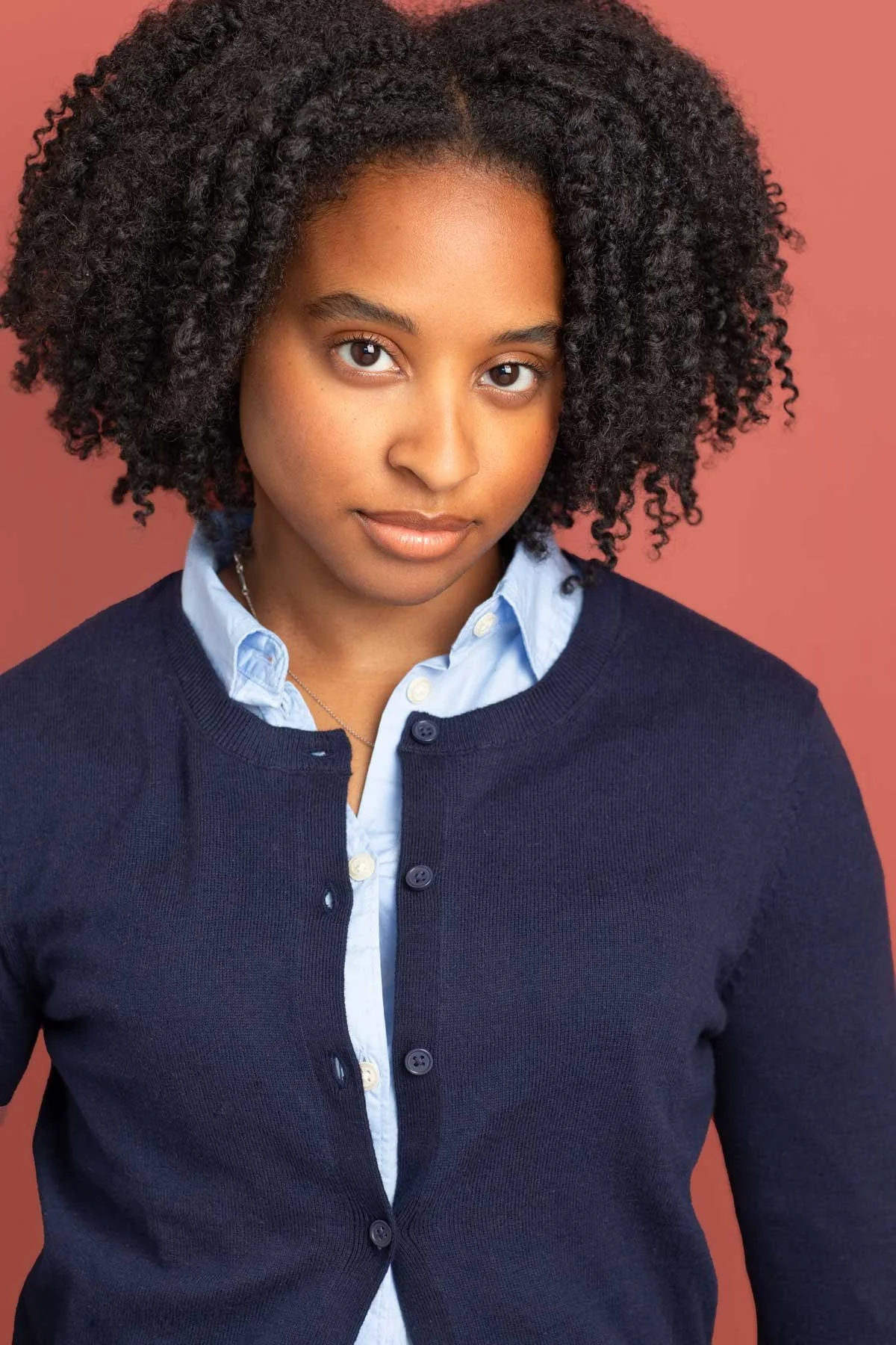 NYC acting headshot of young African American black woman with natural hair dressed in a blue cardigan sweater over a button down shirt, perfect for playing younger teen roles, adult actresses with teen range, shot in Brooklyn studio.
