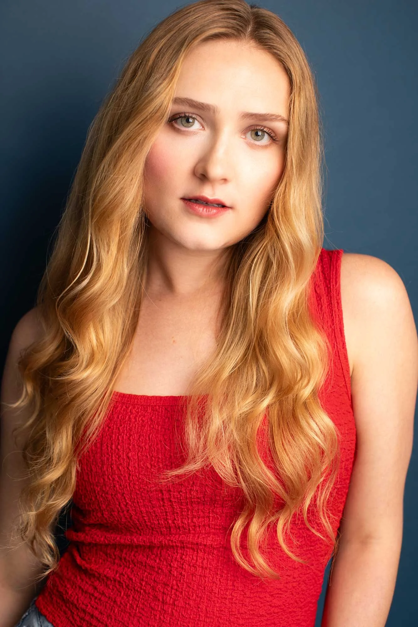 NYC acting headshot of blonde woman in her early 20s with long wavy blonde hair big blue eyes, wearing a red tank top photographed against a dark blue background