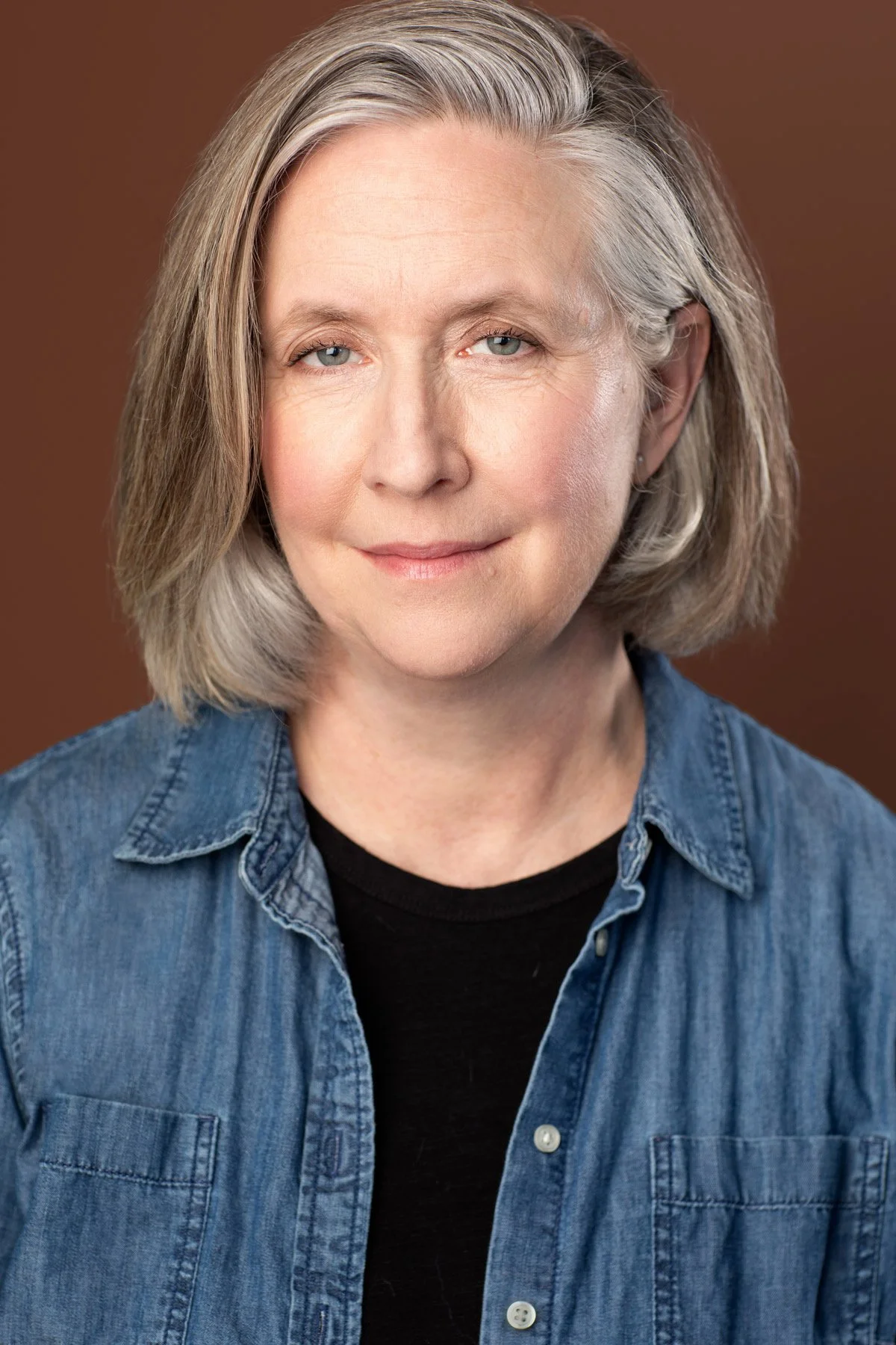 Actor headshot by Hancock Headshots in NYC. A mature actress with shoulder-length gray hair, light eyes, wearing a denim shirt over a black top, smiling softly against a brown background.