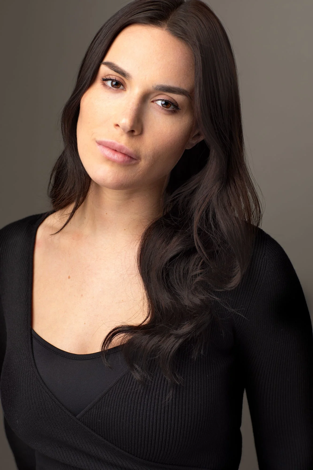 Moody dramatically lit acting headshot of young woman with long dark hair and brown eyes, wearing a black top, photographed in a Brooklyn headshot studio against a charcoal grey background..